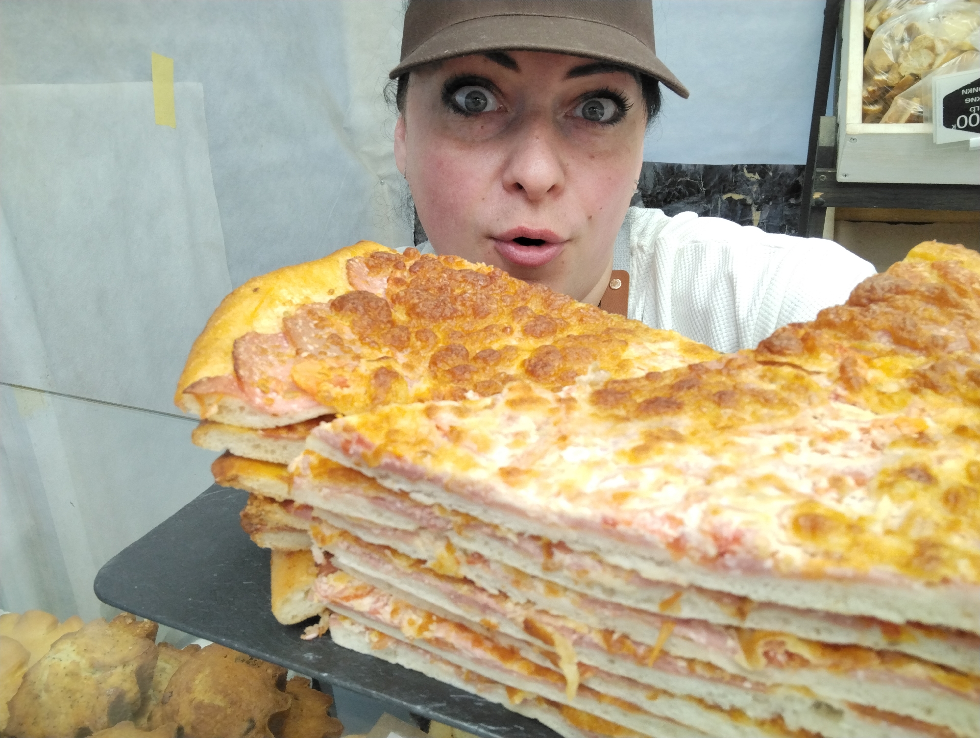 A woman wearing a brown cap is surprised as she presents a tall stack of ham and cheese pizza slices. The pizza sits on a black slate serving tray. The background shows a bakery display case with pastries and a price tag of ".00" on an adjacent shelf. The scene appears to be inside a bakery or food shop. The location of Votkinsk, Russia is not determinable from the visual information.