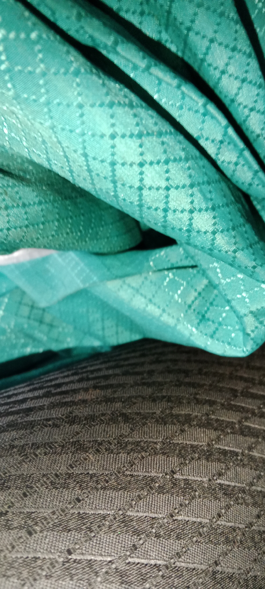 The image shows a close-up, abstract view of folded fabric. The top portion of the image is a teal-colored fabric with a subtle checkered pattern, appearing to be a lighter shade where light hits it directly. There are dark shadows cast by folds in this fabric, creating a sense of depth and texture. Below the teal fabric is a darker, textured material, likely another fabric, with a pattern of horizontal stripes and diamond-like shapes within those stripes. The overall impression is one of layered textiles, with the focus on the patterns and the way light interacts with the folds. There are no people or any clear indication of a specific location within Subang, Indonesia.