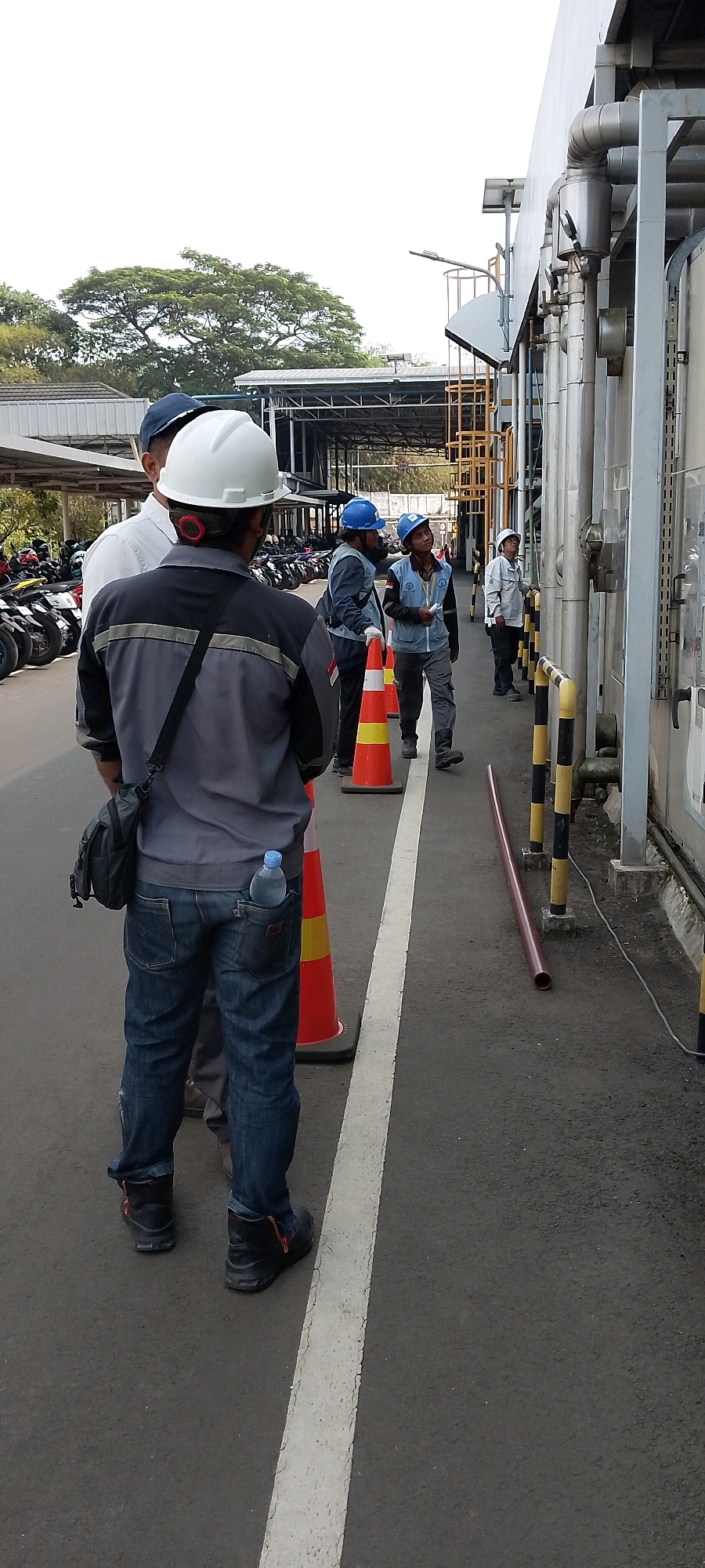In Depok, Indonesia, a group of men in construction attire stand on an asphalt surface marked with a white line, possibly indicating a pathway or zone boundary. Several orange traffic cones with yellow reflective bands are present, some placed in a line. In the background, a building with exposed piping and scaffolding is visible, suggesting an industrial or construction site. The men are wearing hard hats and work clothes, with some appearing to be in discussion. One individual in the foreground has a water bottle tucked into his back pocket. The scene is outdoors, with overcast skies and vegetation visible in the distance, consistent with a semi-urban or industrial setting within Depok.