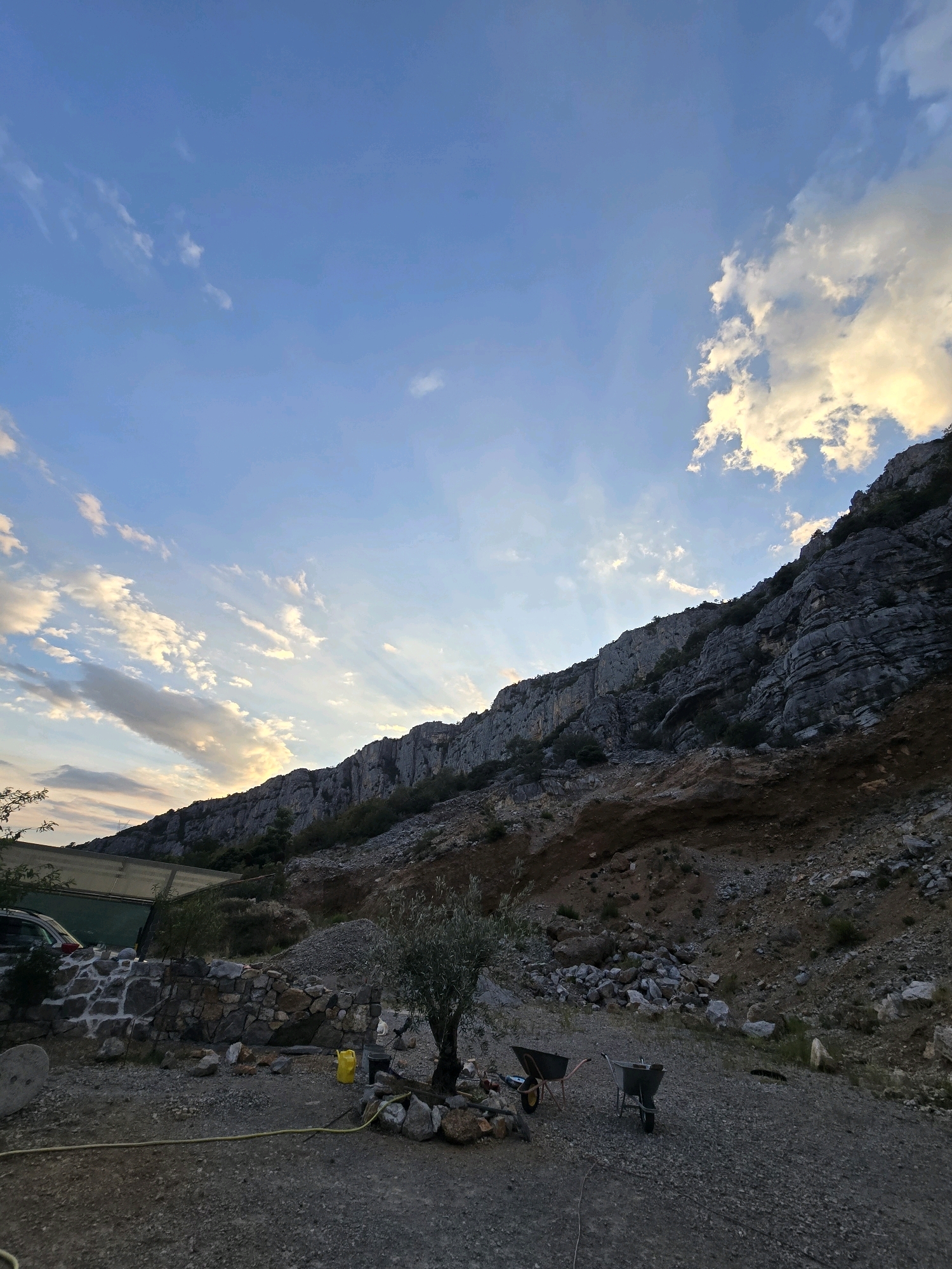 The image depicts an outdoor scene with a mountainous backdrop and a quarry or construction site in the foreground. The sky is a clear blue with scattered white clouds, illuminated by rays of sunlight piercing through them. A large, imposing rock face dominates the upper right portion of the image, with rugged textures and vegetation growing sparsely on its slopes.
In the foreground, a gravelly ground is covered with loose stones and debris. There are two wheelbarrows, one darker and one lighter, positioned on the gravel. To the left, a partially constructed stone wall is visible, with a green tarped structure and a dark-colored car behind it. A small olive tree stands near the center of the frame, surrounded by rocks. A yellow watering can and a black bucket are placed near the base of the tree. A yellow hose lies on the ground, extending across the lower part of the image.
The scene suggests a working quarry or a construction project in a mountainous area. The lighting indicates it is either early morning or late afternoon, with the sun casting long shadows. The location is identified as Podgorica, Montenegro. There are no people visible in the image.