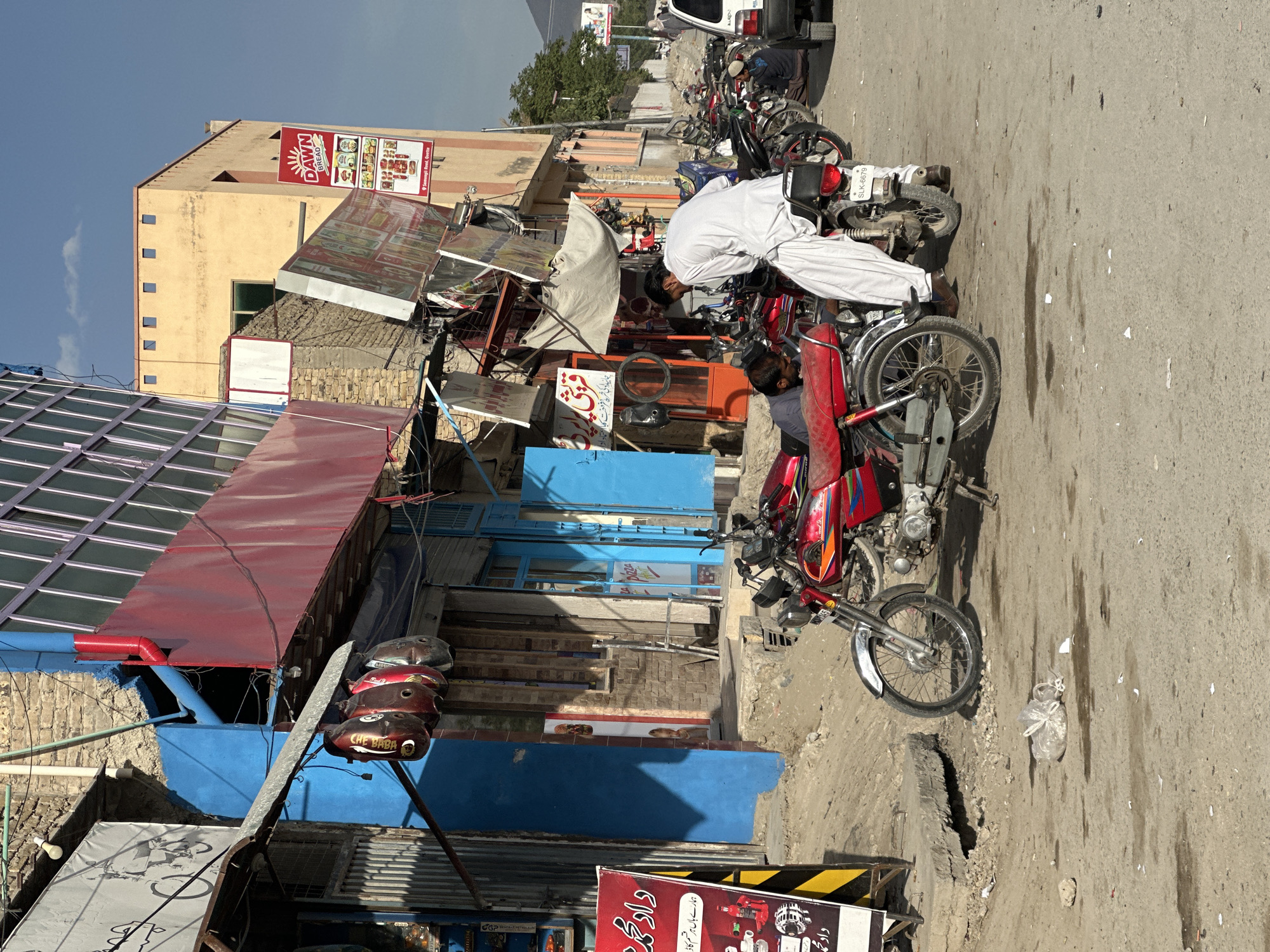 The image depicts a street scene in Quetta City Tehsil, Pakistan. A man in traditional white clothing is bent over a red motorcycle parked on the street. Several other motorcycles are parked nearby. Shops and buildings line the street, with signs and awnings visible. The "DAWN BREAD" sign and the "CHE BABA" label are distinct elements. The road is paved, and there's some litter present, including a plastic bag. The buildings are a mix of stone and painted structures.