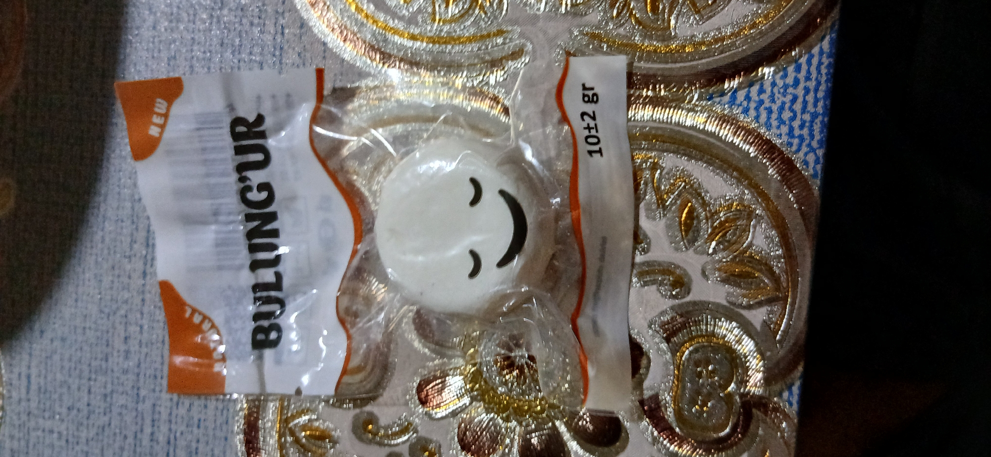 The image shows a close-up of a small, white, round object with a smiling face printed on it, enclosed in a clear plastic wrapper. The wrapper has some text printed on it, including "BULUNG'UR" and "NEW". The object appears to be a toy or a candy. The background is a patterned fabric, possibly a tablecloth or upholstery. There is no indication of the city of Andijani, Uzbekistan, in the image. The scene is static and does not depict any actions or interactions.