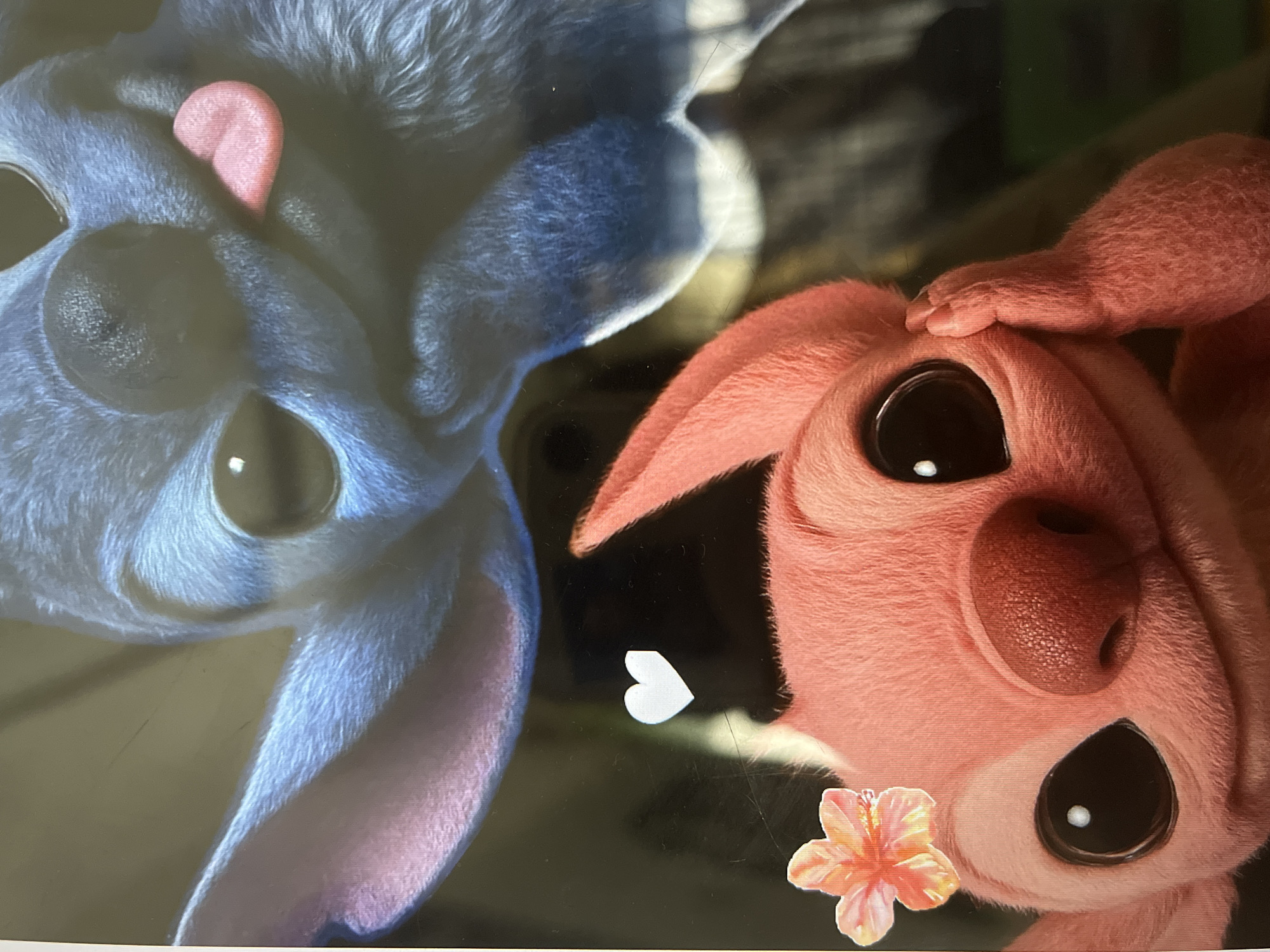 The image is a close-up of two cartoon characters. The character in the foreground is pink with large, dark eyes, a red nose, and a hand resting on its face. It is adorned with a small orange and yellow flower on its head. The character in the background is blue, with large ears and its tongue sticking out. A small white heart is visible between the two characters. The background is dark and out of focus.