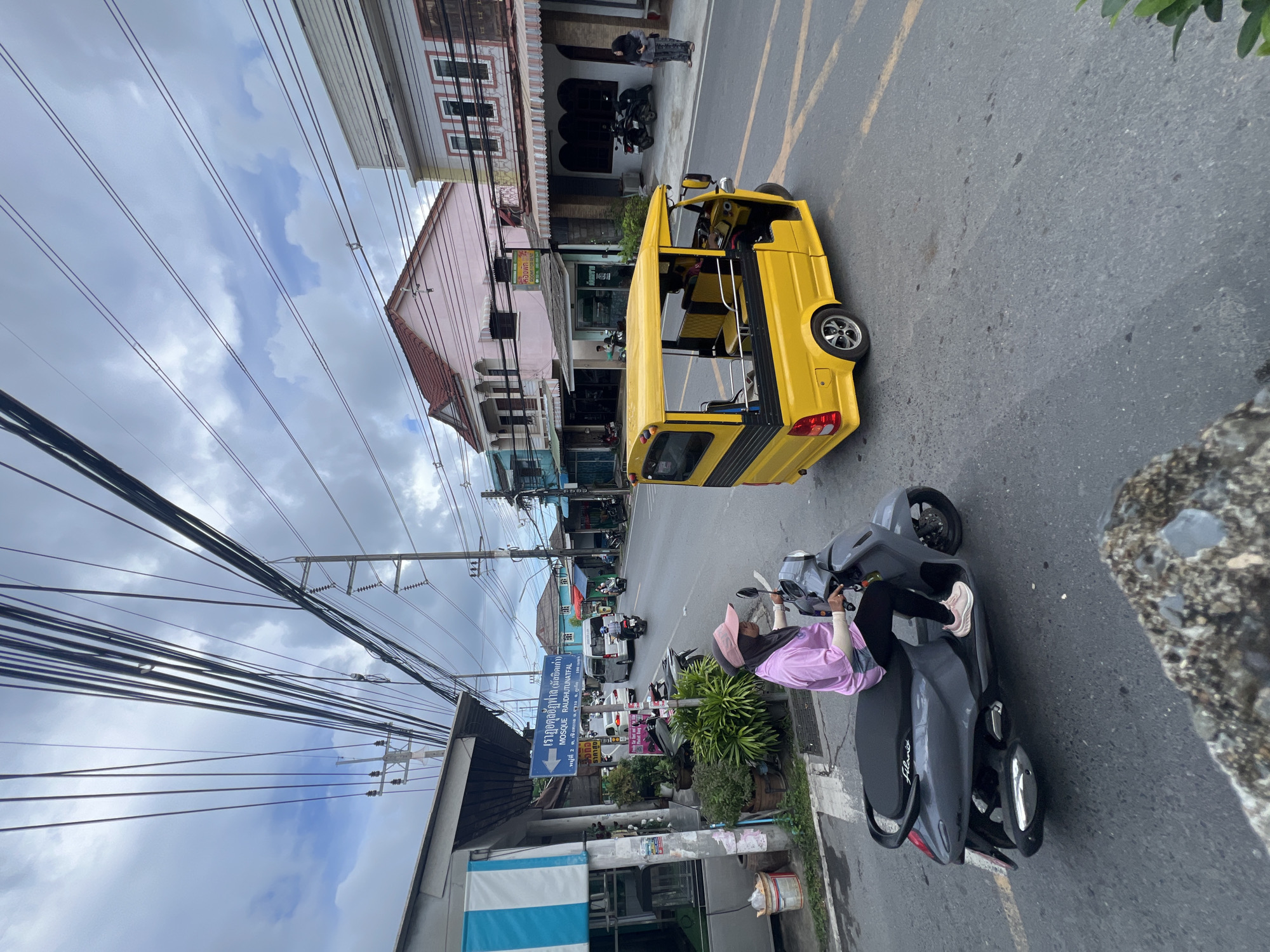 In Choeng Thale, Thailand, a person wearing a pink long-sleeved shirt and a pink headscarf rides a dark gray scooter on an asphalt road. The scooter has a black seat and a silver exhaust pipe. Behind the scooter, a bright yellow tuk-tuk with black trim and yellow seating is driving in the same direction. The tuk-tuk has an open passenger area and a driver visible. The road is marked with yellow lines.
On the left side of the road, there is a building with a blue and white striped awning and a blue directional sign pointing left, indicating "MOSQUE RAUDHUTUNAFATAL" and a distance of "100 meters." Several potted plants are situated near the building. Further down the street, other buildings are visible, including a pink building with a red roof and a light-colored building with multiple windows. There are also power lines overhead, supported by utility poles. Several other motorcycles and a white van are present on the road in the distance. A person in a gray headscarf and gray clothing is standing on the right sidewalk near the light-colored building. The sky is cloudy.