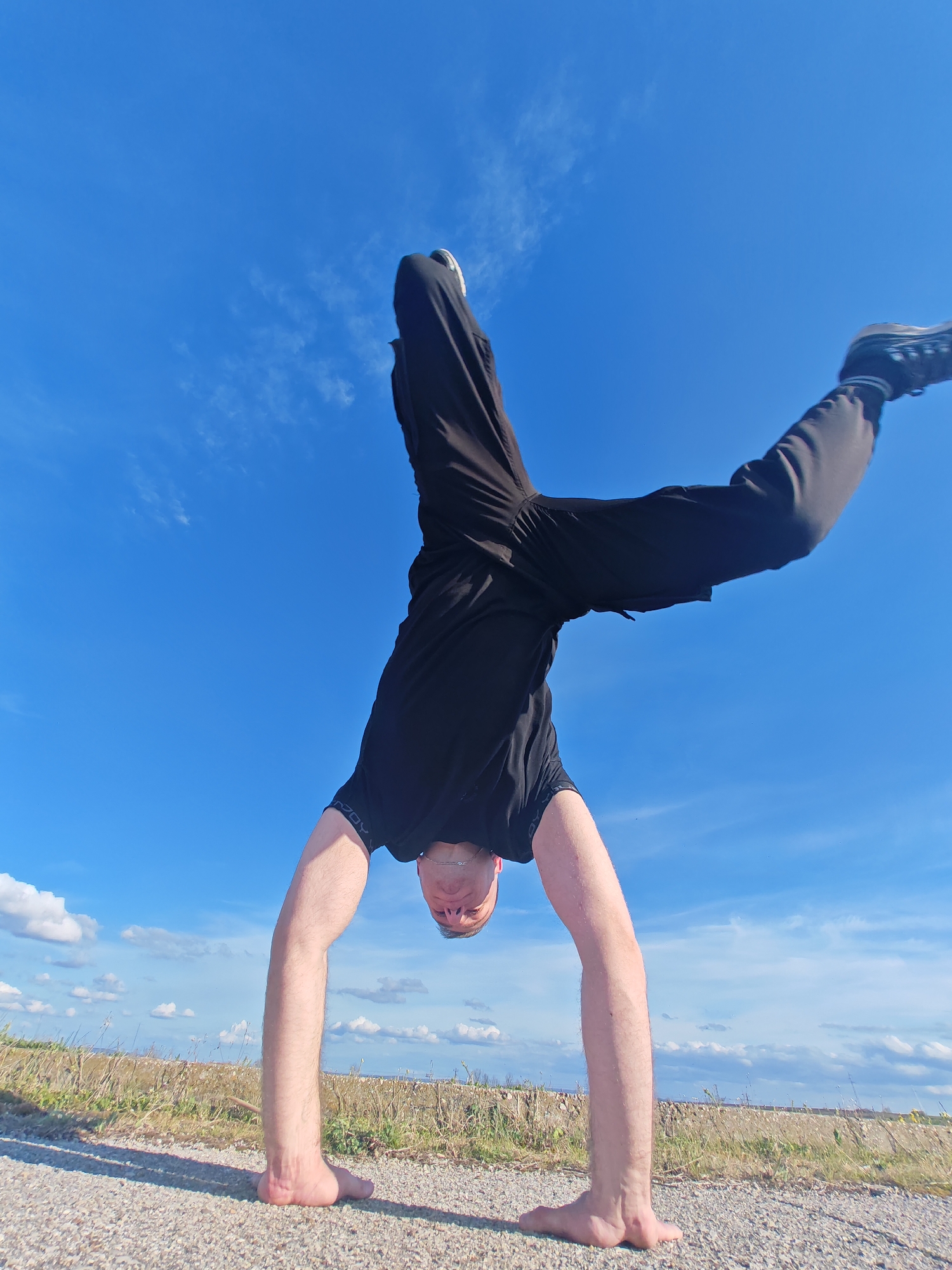 A person is performing a handstand on a paved surface. The person is wearing black pants and a black t-shirt, and is barefoot with the exception of the right foot which has a black shoe. The background shows a field with dry grass, and a bright blue sky with some clouds. The location is likely in a rural area of Shumen, Bulgaria, as there is no urban landscape visible.