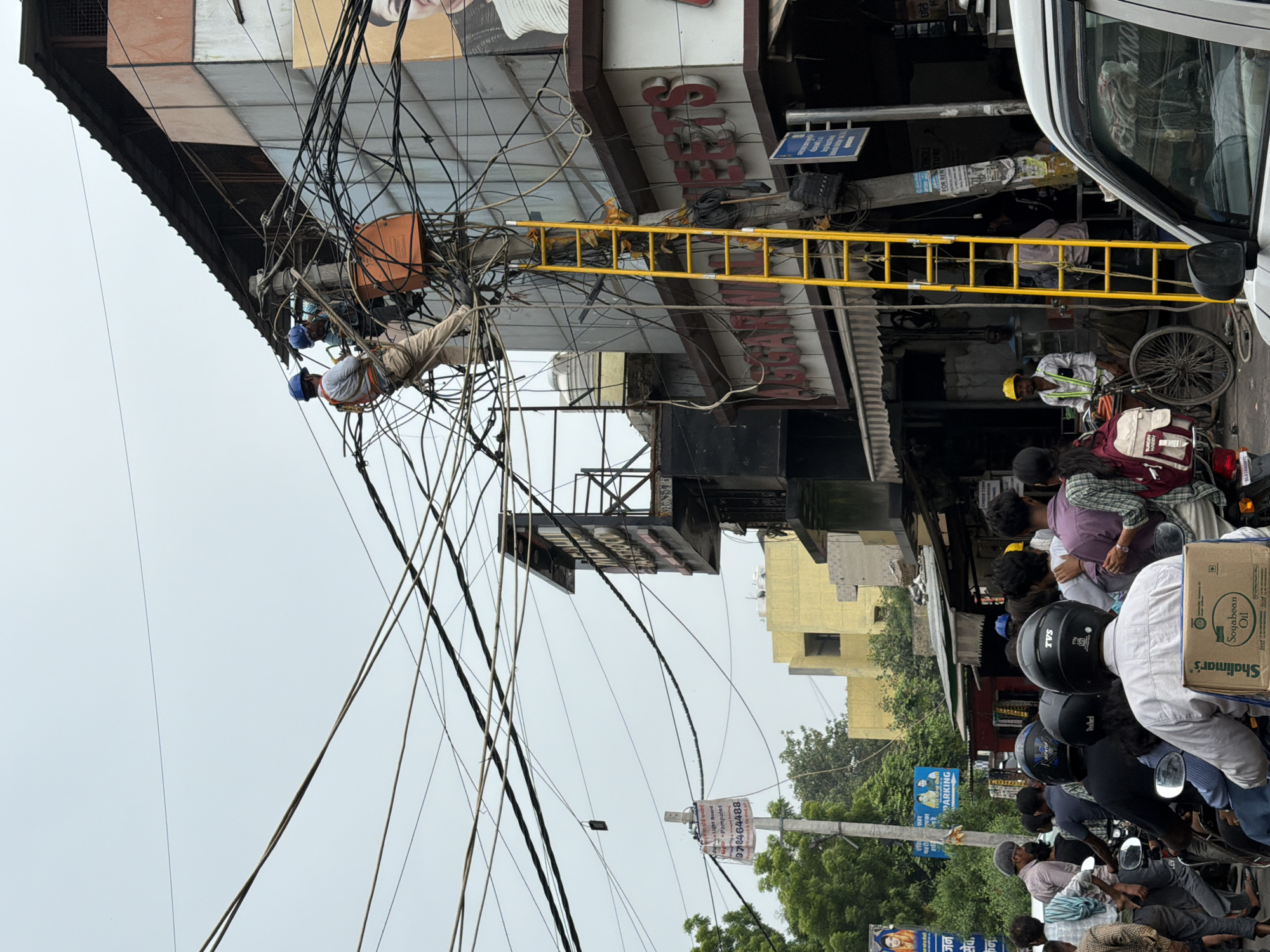 This image depicts a street scene in Delhi, India, likely a market or commercial area given the shops visible. In the upper portion of the image, two individuals, presumably electricians, are working on a dense cluster of overhead wires. They are wearing safety harnesses and helmets. One is positioned on a utility pole, while the other is on a tall, bright yellow ladder that extends up to the wires. The background features multi-story buildings, one with a faded advertisement featuring a person's face. Below the utility work, the street is bustling with activity. Several people are on motorcycles and scooters, suggesting moderate traffic. Some individuals are walking, and there are storefronts lining the street. A man in a yellow hard hat stands near a bicycle, observing the scene. The overall impression is one of urban life with ongoing infrastructure maintenance.