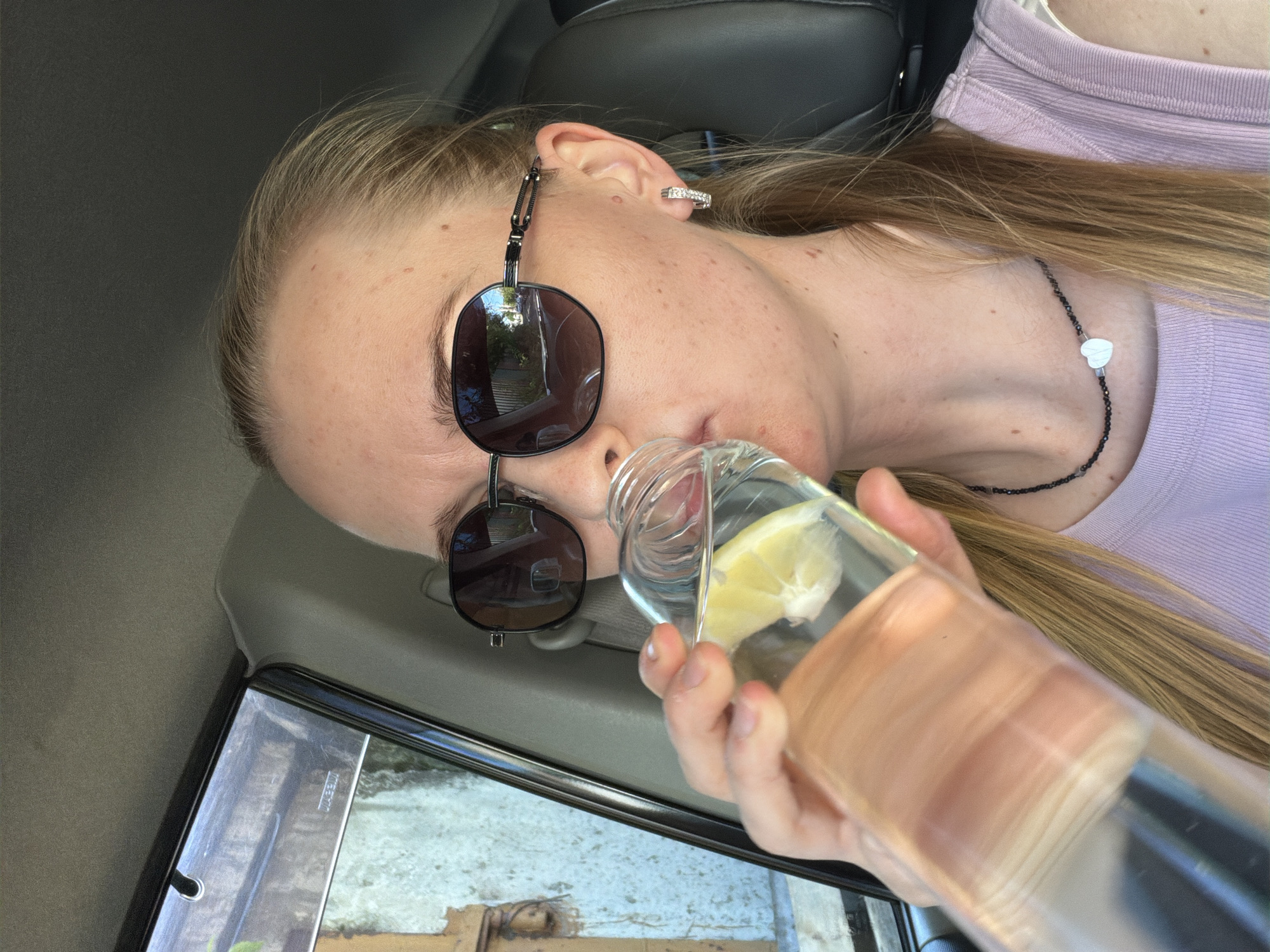 A woman with blonde hair pulled back, wearing sunglasses and a light purple tank top, is sitting in the passenger seat of a vehicle. She is holding a glass bottle filled with water and a lemon slice and is in the process of drinking from it. Her right hand is visible, holding the bottle. She is wearing a necklace with a white heart pendant and hoop earrings with small stones. The interior of the car is visible, including the headrest and part of the window. The outdoor environment is reflected in the sunglasses.