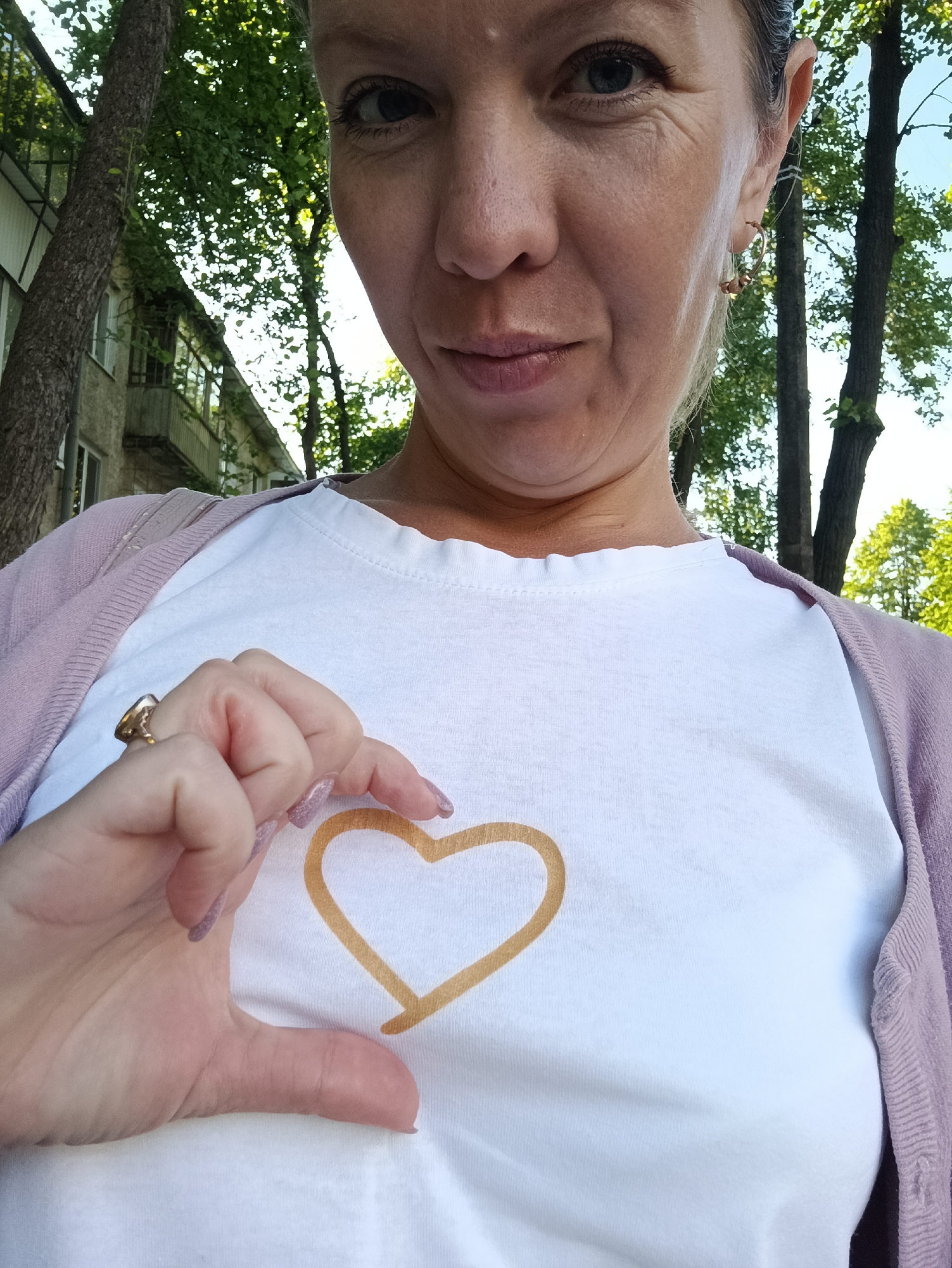 A woman with blonde hair, wearing a white t-shirt with a gold heart drawn on it and a light purple cardigan, is holding her hand to form half of a heart shape in front of the t-shirt. She is outdoors, with a tree and a building visible in the background, suggesting an urban or suburban setting. The image appears to have been taken on a sunny day. The location is identified as Chaikovsky, Russia.
