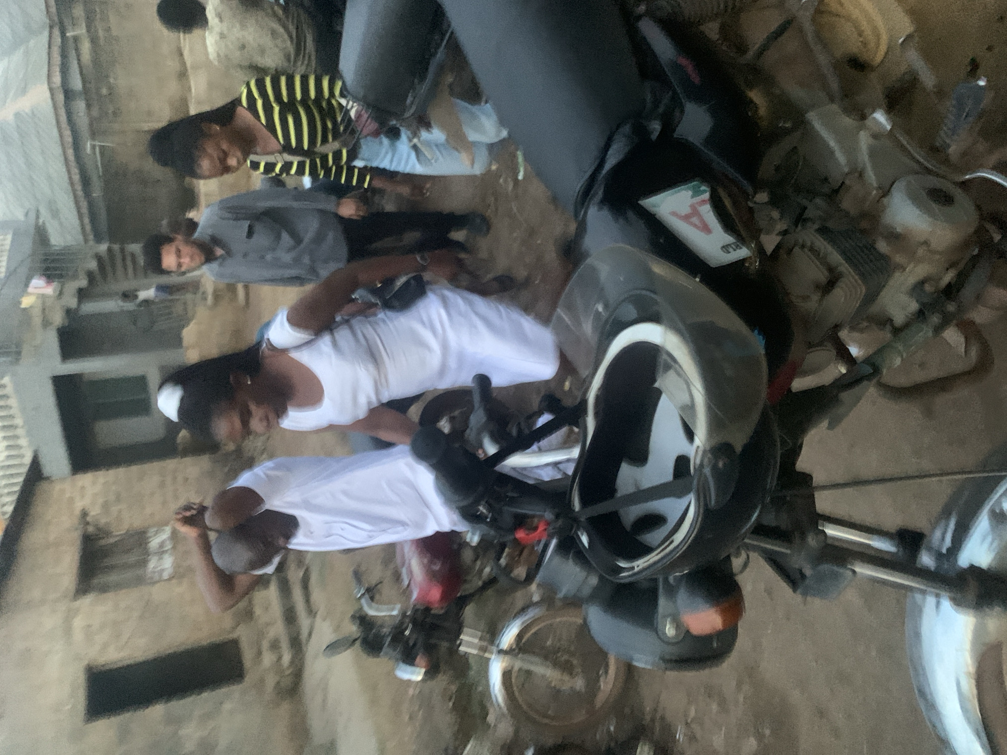 In Shagamu, Nigeria, an outdoor scene unfolds with several individuals gathered around motorcycles. A man in a white t-shirt is in the foreground, his arms raised to his head. Beside him, a woman in a white dress stands, her gaze directed downwards. Behind them, a man in a grey button-down shirt and black pants walks away from the camera. To his right, a woman in a yellow and black striped long-sleeved shirt and light blue jeans stands with her head bowed. A motorcycle with a red fuel tank and a black seat is parked to the left, while another motorcycle with a black seat and a helmet on its handlebars is in the immediate foreground. The ground is dirt, and in the background, buildings with rough textures are visible, suggesting a residential or urban environment.