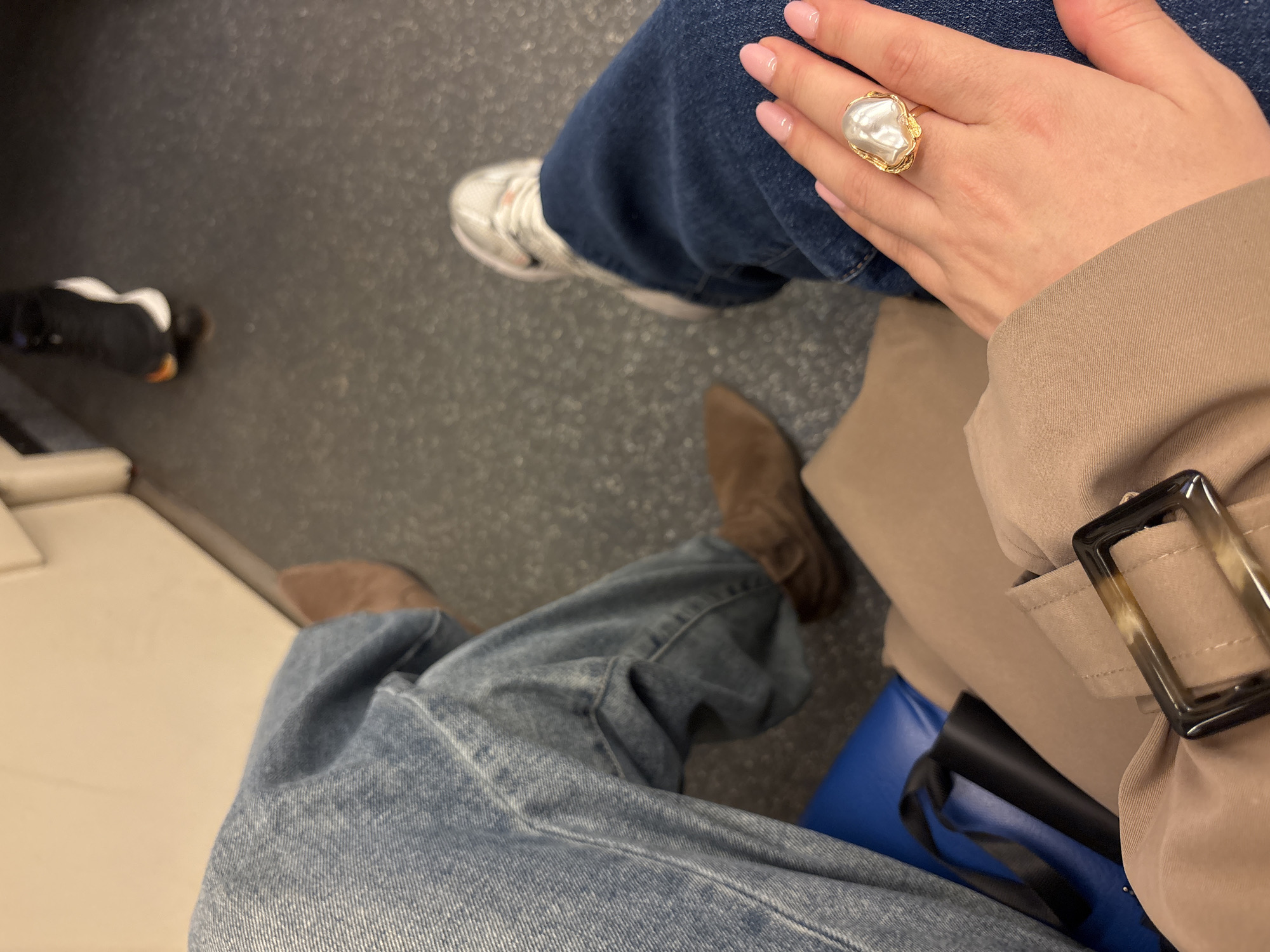 The image shows two seated individuals on what appears to be public transportation, possibly a train, based on the glimpse of an adjacent white panel and the overall setting. The individual on the right is wearing a beige trench coat with a distinctive buckle detail. They have a large, ornamental ring on their finger. They are wearing jeans and white sneakers. The individual on the left is wearing jeans and brown boots. The ground is dark and looks like metal. A blue bag is partially visible beneath the trench coat. There is no explicit indication of Moscow or Russia in the image.