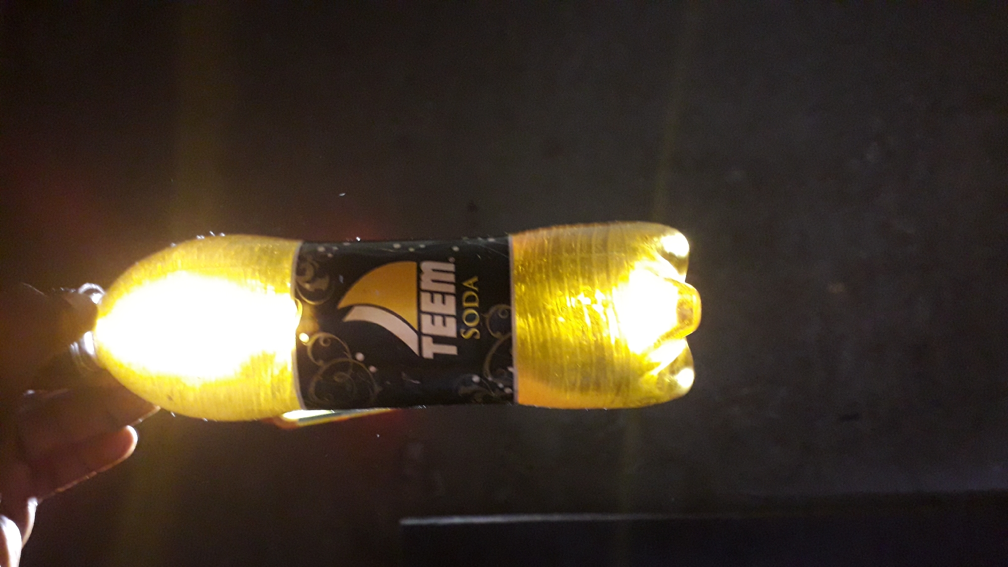 A bottle of "TEEM Soda" is brightly illuminated, obscuring details of its yellow contents and plastic structure. The bottle is held by a hand on the left. The background is a dark, undefined area. Nkpor, Nigeria is not discernable from the image.