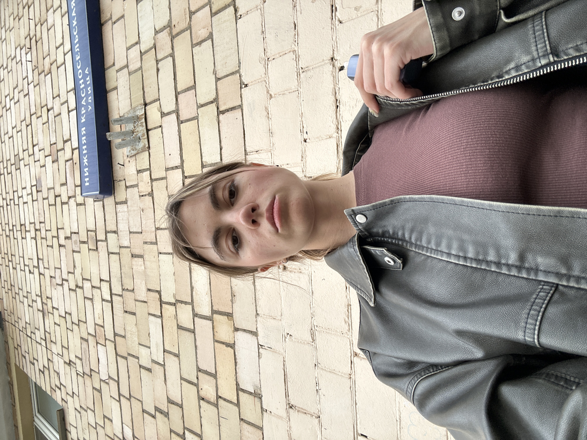 A low-angle, medium shot captures a young woman from the chest up, standing in front of a light-colored brick building in Moscow, Russia. She is wearing a dark leather jacket over a maroon ribbed top. Her hand, holding a blue object, is near the zipper of her jacket. Above and behind her, a street sign is visible on the brick wall, reading "Нижняя Красносельская улица" (Nizhnyaya Krasnoselskaya Street). The building appears to be an apartment block, with a window visible in the upper left corner of the frame. The lighting suggests an overcast day.