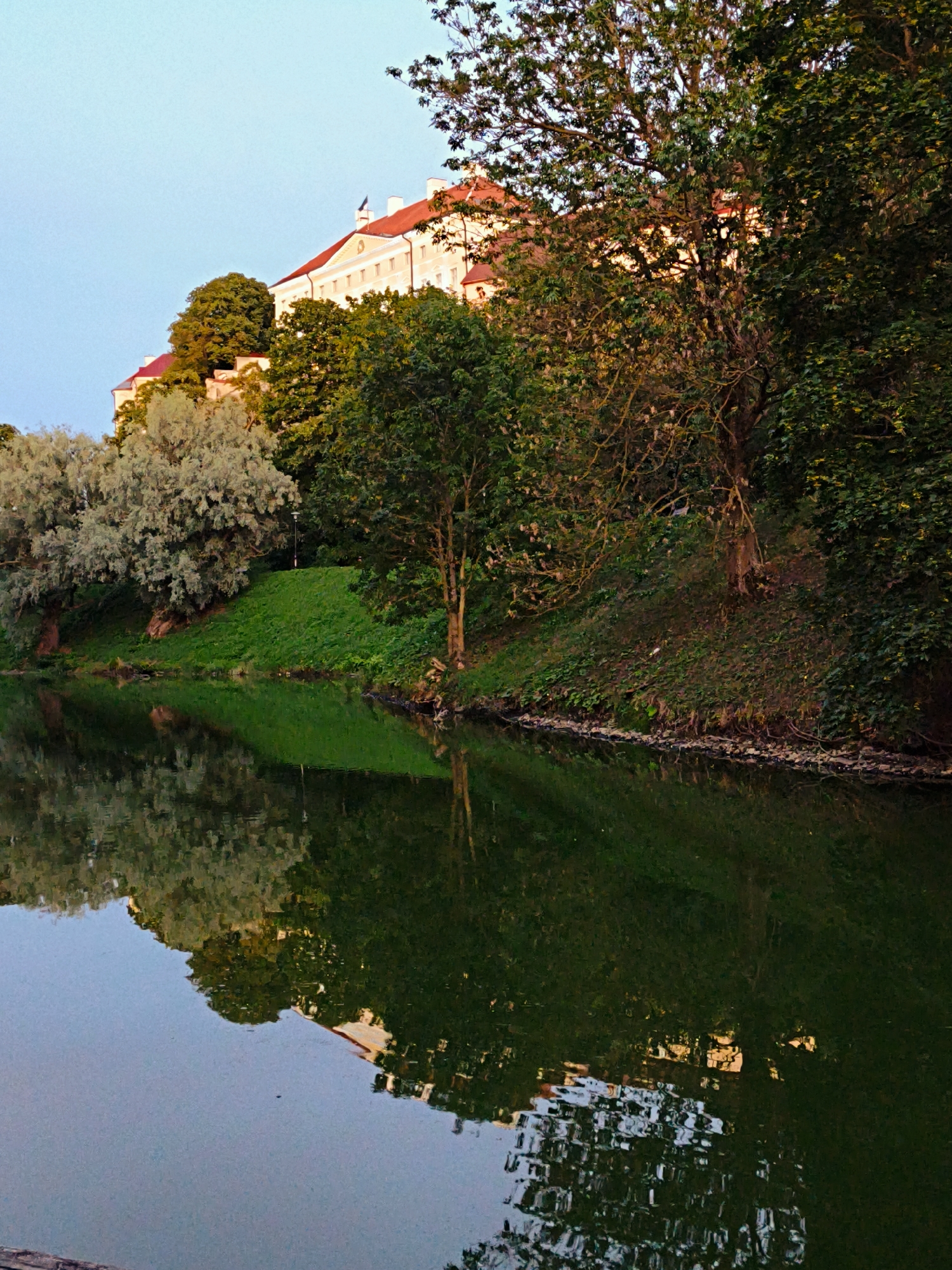 The image depicts a tranquil waterside scene in Tallinn, Estonia, with a historical building situated atop a hill. The building, characterized by its light-colored facade and red-tiled roof, is partially obscured by lush green trees. The foreground features a body of water, likely a pond or canal, reflecting the surrounding greenery and the building itself. The water's surface is disturbed by subtle ripples, creating an abstract pattern of reflected light and color. The vegetation appears dense, with a mix of deciduous trees and possibly some shrubs, contributing to the natural beauty of the setting. The sky above is a soft gradient of pale blue, suggesting either early morning or late evening light. The overall atmosphere of the image is serene and picturesque, capturing a harmonious blend of nature and architecture in an urban environment.