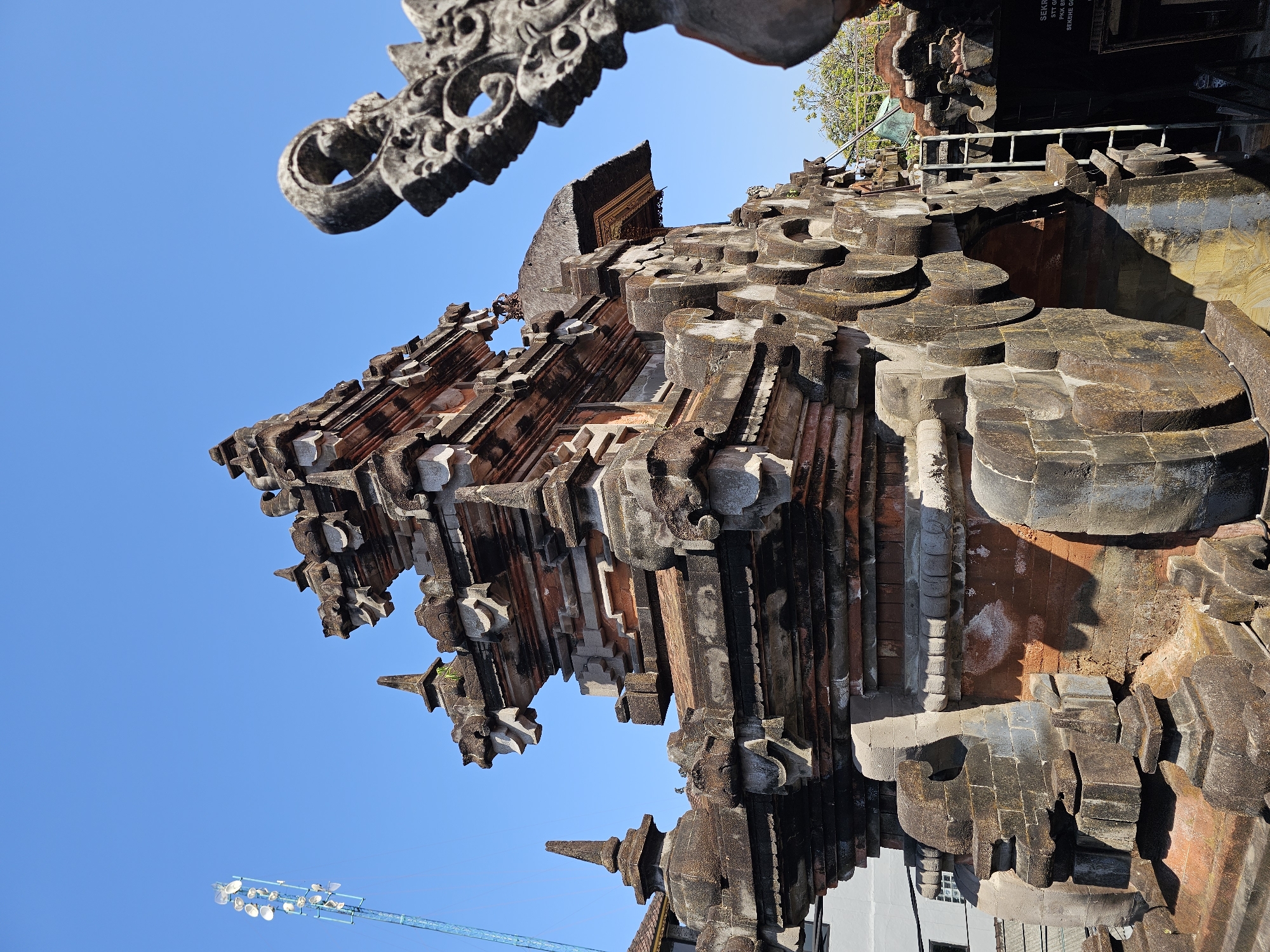 An ornate, multi-tiered stone structure, likely a temple gate or pavilion, stands against a clear blue sky. The structure is made of weathered, reddish-brown and grey stone, with intricate carvings and architectural details, including what appear to be dragon or mythical creature heads adorning the upper levels. The stone shows signs of age and has a mossy or discolored patina in places. In the foreground and right side of the image, parts of another carved stone element are visible, also weathered and detailed. Behind the main structure, a modern-looking white building and a tall telecommunications tower with multiple antennas are visible, contrasting with the traditional architecture. A metal ladder leans against a section of the stone structure on the right side, suggesting ongoing work or maintenance. The scene appears to be outdoors, under bright sunlight, casting distinct shadows. The text on a sign near the bottom right corner is partially visible and in Indonesian, suggesting the location is in Indonesia. Based on the architectural style and the presence of Indonesian text, this structure is likely in Denpasar, Indonesia, possibly a religious site.