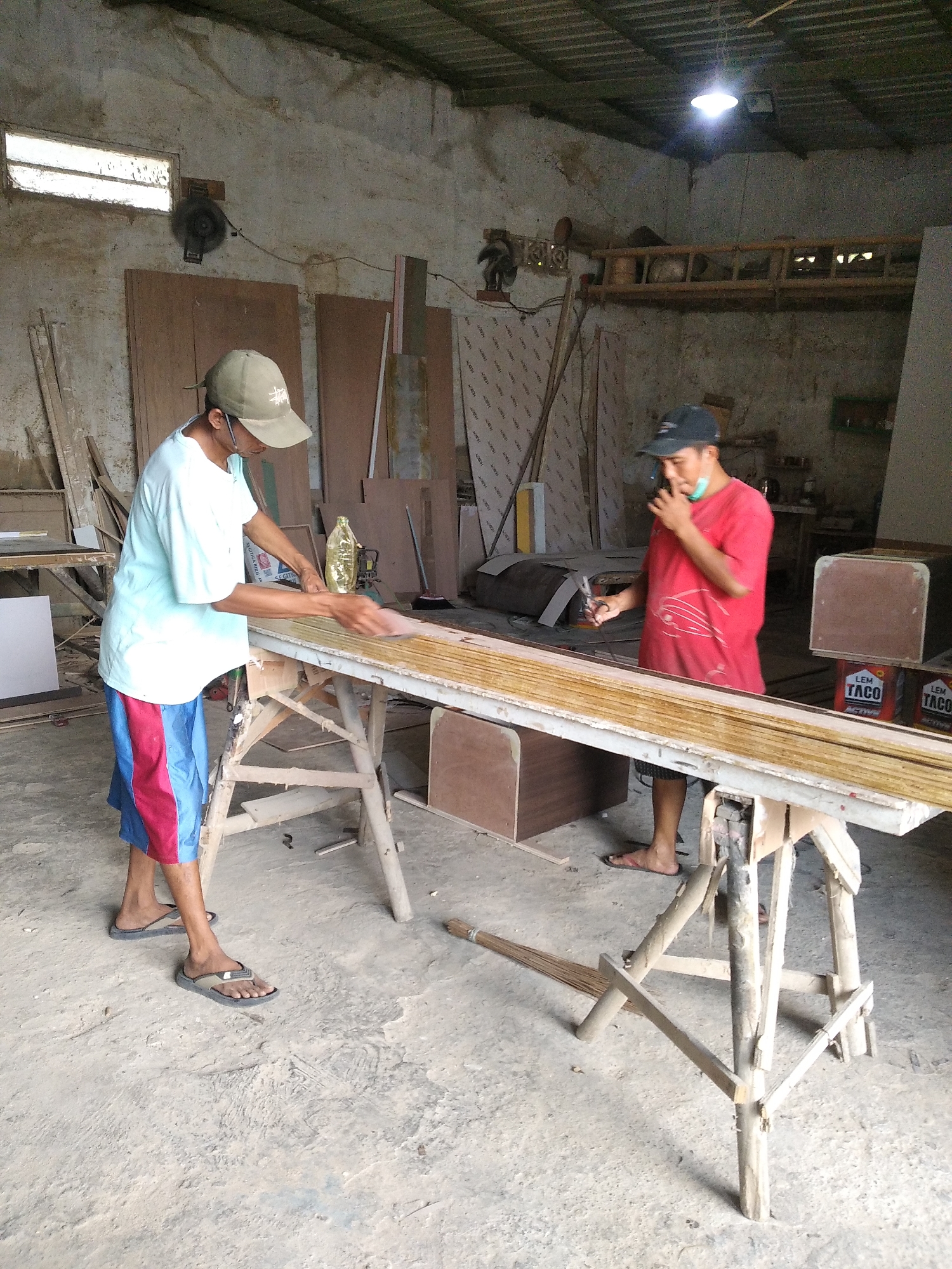 In a workshop located in Jumerto, Indonesia, two men are engaged in woodworking. The man on the left, wearing a light blue t-shirt, red and blue shorts, and a green cap, is sanding a stack of wooden planks supported by a wooden sawhorse. He is holding a sanding block in his right hand and appears focused on his task. To his right, a man in a red t-shirt and black cap is standing near the end of the wooden planks, holding a tool, possibly pliers, in his right hand and touching his face with his left hand, as if taking a break or contemplating his next move.

The workshop appears to be constructed with a metal roof and rough concrete or dirt flooring. The walls are unfinished concrete, with some wooden panels and construction materials visible in the background. Stacks of wood, tools, and containers are scattered around the space, indicating an active workspace. A bright fluorescent light hangs from the ceiling, illuminating the scene. Two buckets labeled "LEM TACO" are visible near the end of the planks. The overall impression is one of a functional, albeit somewhat rustic, woodworking facility.