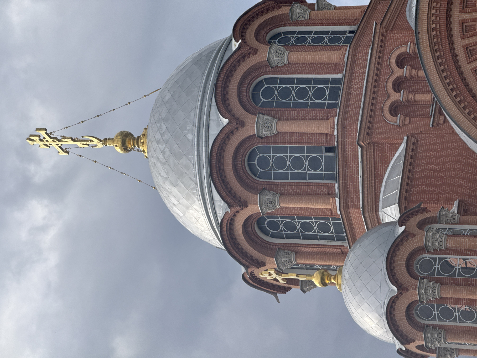 A low-angle shot shows the dome and brickwork of a building, presumably a church or religious structure, against a cloudy sky in Unknown, Unknown. The dome is silver and possibly metallic in appearance. Atop the dome is a gold cross, secured by chains. The building's facade consists of red brick with arched windows. Columns with decorative capitals are visible between the windows. The scene's lighting is diffused due to the overcast sky.