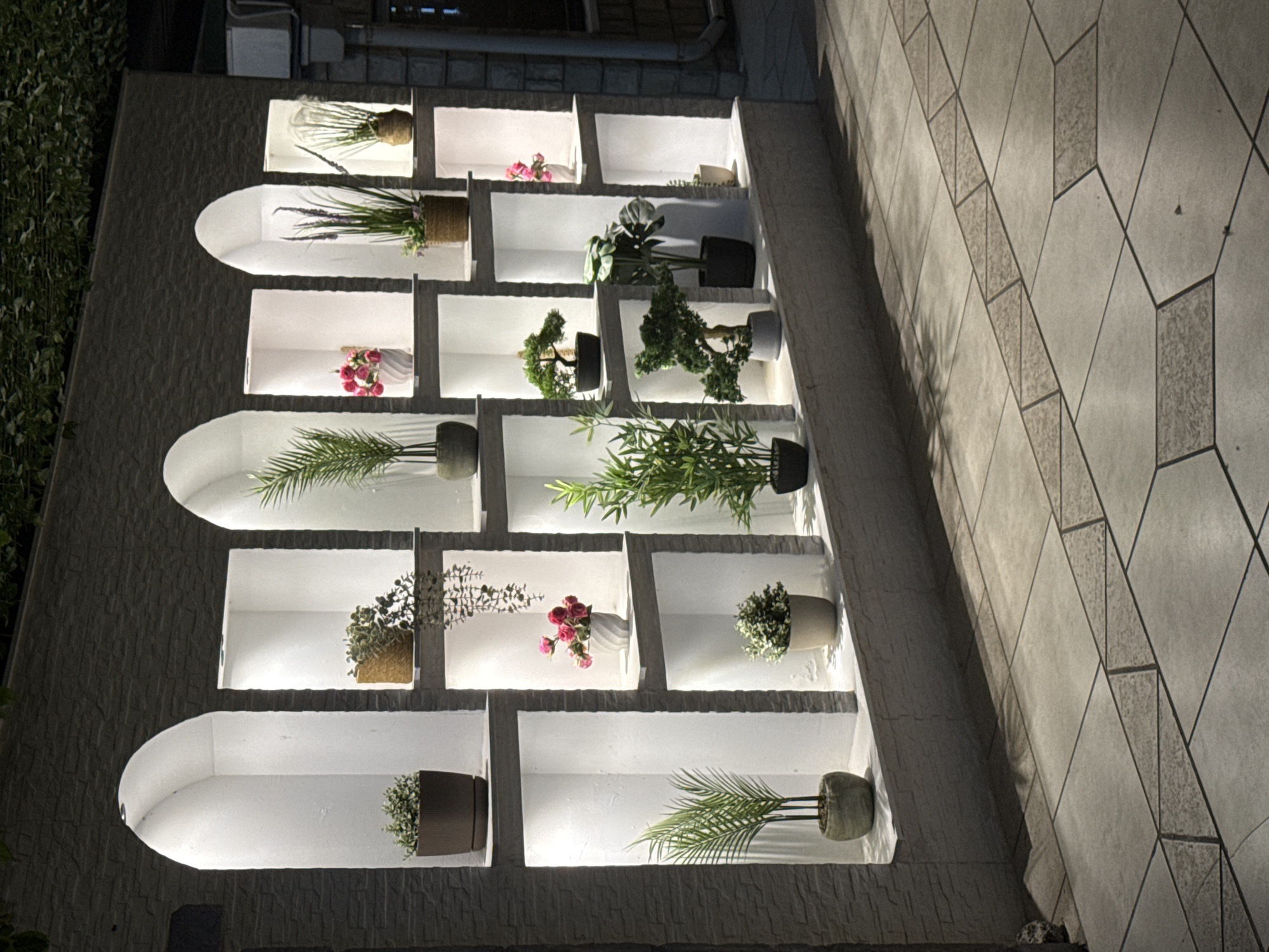 An outdoor display of potted plants is illuminated by spotlights in recessed niches along a textured stone wall. The display is positioned on a tiled patio, with geometric patterns in the tiling. The scene is likely in Budva, Montenegro.