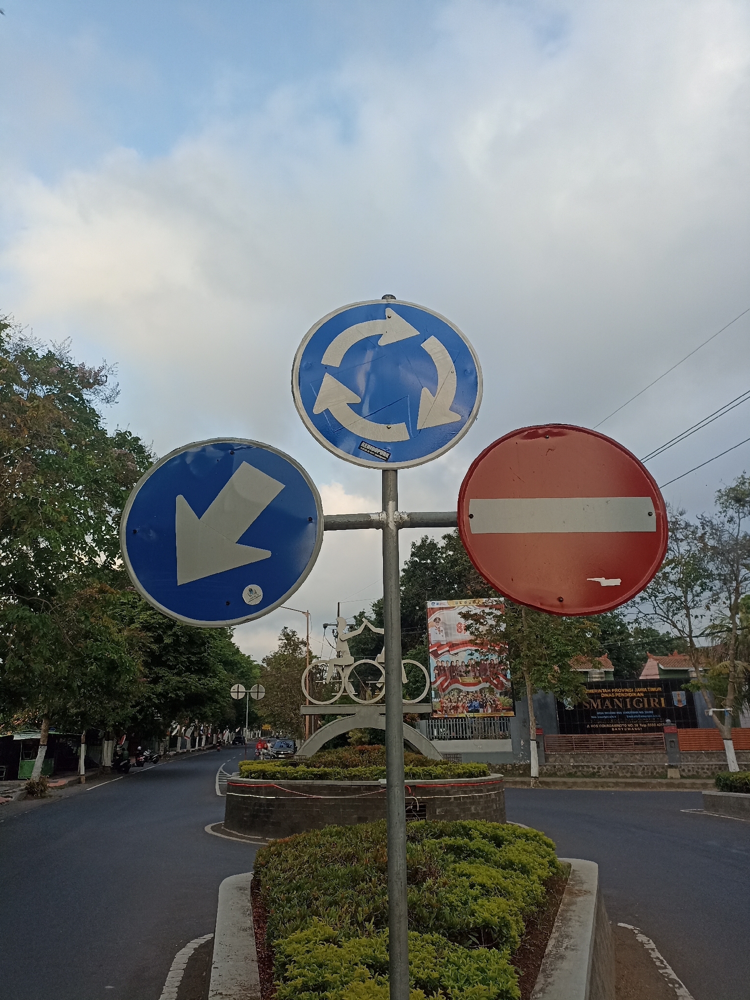 An eye-level, medium shot captures a traffic signpost with three signs against a cloudy sky at the intersection of a paved road in Mojopanggung, Indonesia. The signpost, a vertical metal pole, supports three circular signs. The top sign is blue with a white border and depicts three white arrows forming a circular path, indicating a roundabout. Below it, to the left, is another blue sign with a white border, featuring a large white arrow pointing downwards and slightly to the left. To the right of the roundabout sign is a red and white sign, a circle with a white horizontal bar, signifying "Do Not Enter" or "No Entry".
In the background, the road curves to the left, lined with trees and some parked motorcycles. Further in the distance, a monument with a bicycle sculpture is visible, and beyond that, buildings and a large banner for "SMAN GIRI". The road is dark asphalt, with white markings. A concrete planter box filled with green bushes is in the foreground, surrounding the base of the signpost. The sky is predominantly light blue with scattered white clouds. The scene appears to be during daylight, likely late afternoon given the soft lighting. No people are clearly identifiable in the foreground, though some individuals and vehicles are faintly visible on the road in the background.