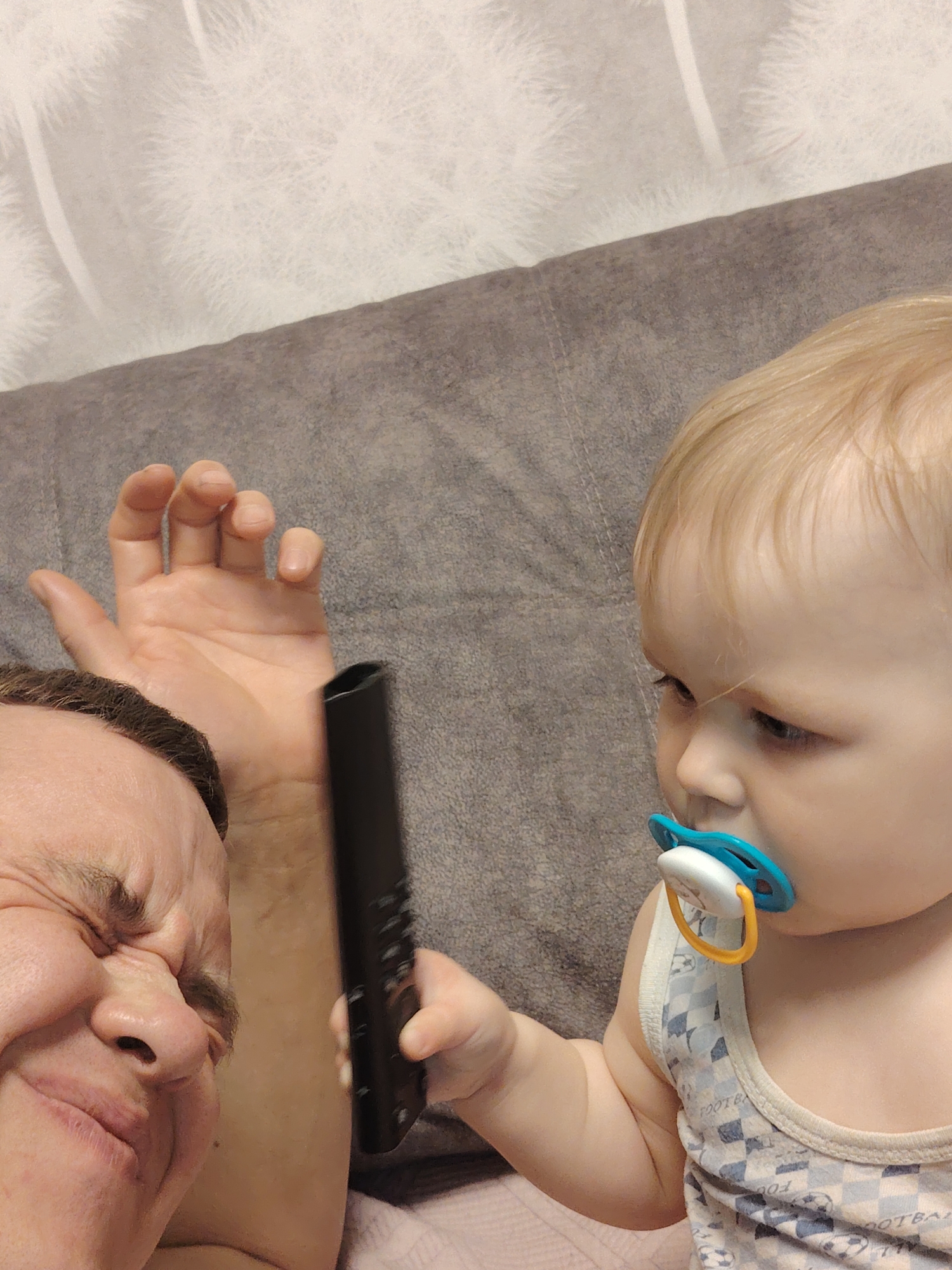 A close-up shot shows a man and a baby. The man is visible from the left, his face scrunched up with his eyes closed. He has short dark hair and is partially lying down, as his arm is also visible. The baby, wearing a tank top with a checkered pattern and soccer balls, is holding a black remote control. The baby also has a pacifier in its mouth and is looking at the remote. The background consists of a gray pillow and a white patterned blanket. The image was taken in Chaikovsky, Russia.