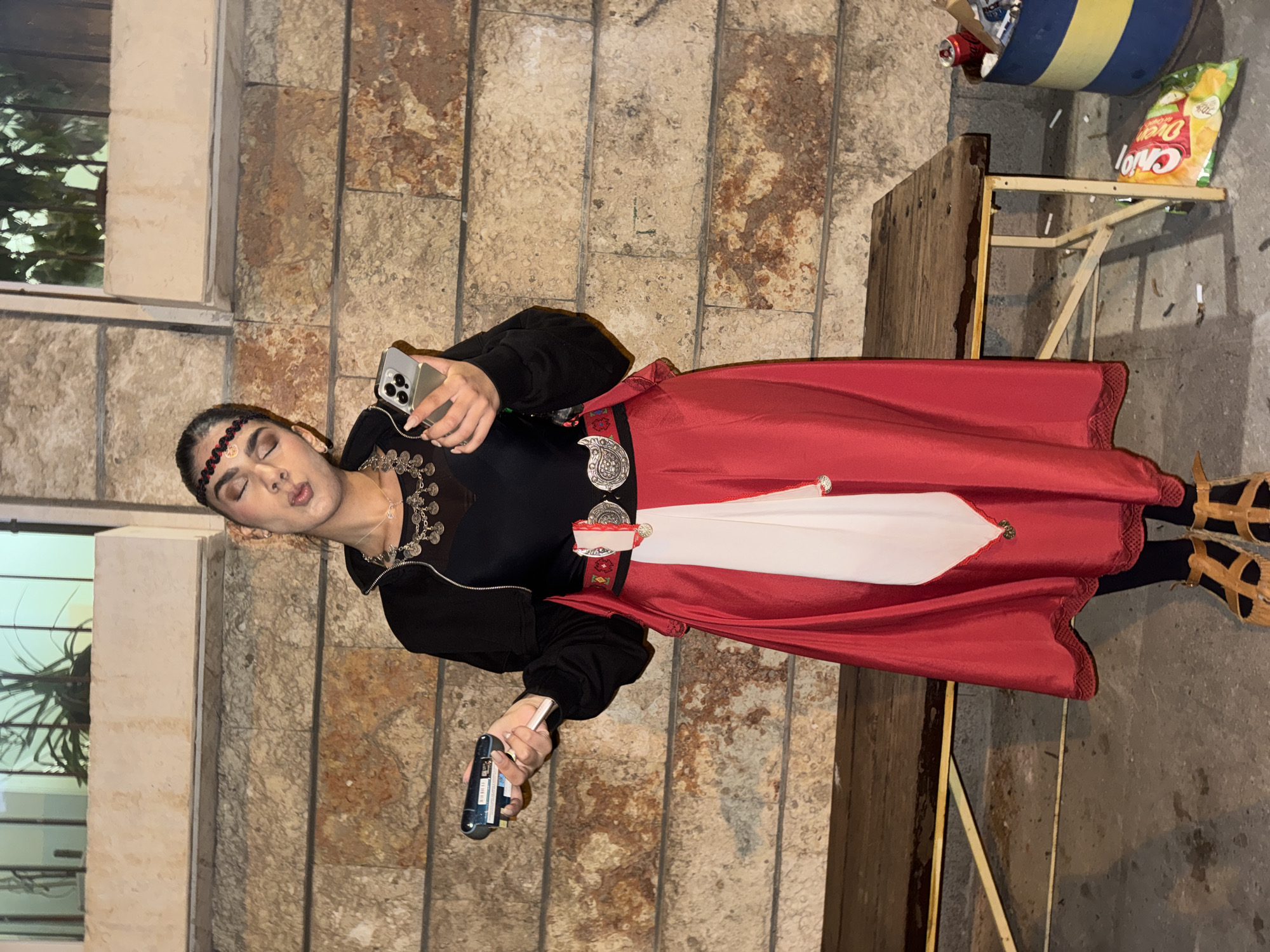 A person is standing in front of a stone wall in an urban environment. The person is holding a smartphone in their left hand and an electronic device in their right. The person is wearing a black jacket, black shirt with a silver necklace, a red skirt with a white design, a decorative belt, and a headband. Next to the person is a wooden table with a bag of chips and a container on top. The city and state of Unknown, Unknown cannot be verified based on the visual data.