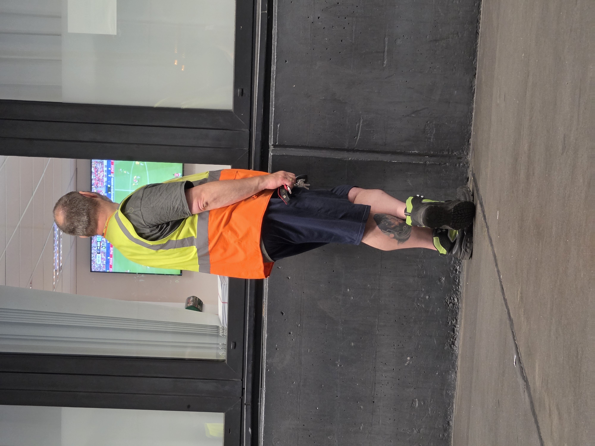 A man wearing a high-visibility vest over a gray t-shirt and dark blue shorts is standing in front of a gray concrete wall, looking at a television screen displaying a soccer match. The man has tattoos on his left calf and is wearing dark boots with bright green accents. He is holding a set of keys in his right hand. Behind him, a large window reveals a glimpse of an interior space with vertical blinds and office lighting. The scene appears to be outdoors, possibly near a stadium or sports facility. There is no identifiable information suggesting the location is in Frankfurt, Germany.