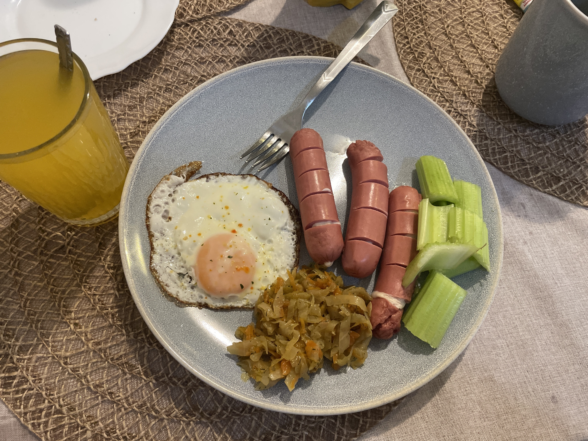 A plate of food is on a woven placemat on a tan tablecloth. The plate contains a fried egg, three sausages cut with incisions, cooked shredded cabbage, and cut celery stalks. A fork rests on the plate. A glass of orange juice is to the left of the plate, and a white plate is partially visible in the upper left corner. A grey mug is to the right of the plate. The scene suggests a meal setting indoors, possibly in Bar, Montenegro. No people are directly visible.