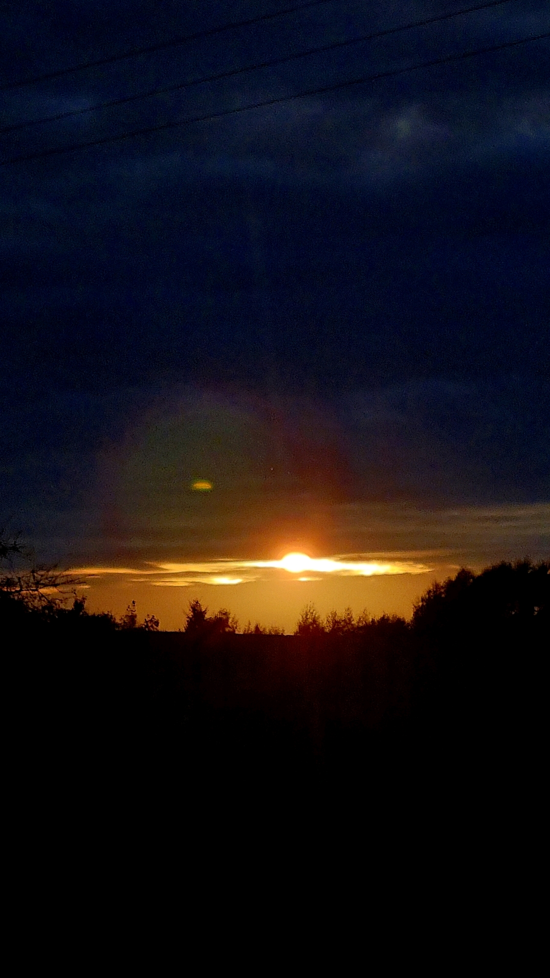 A sunset is depicted over a silhouetted tree line, with a dark, cloudy sky above. The sun is a bright, orange disc partially obscured by clouds, casting a warm glow across the horizon. A faint rainbow-like arc is visible in the sky to the left of the sun, and a small, bright circular lens flare is present above and to the left of the sun. In the foreground, the dark silhouettes of trees and foliage stretch across the bottom of the frame. Power lines are faintly visible in the upper portion of the image. The location is noted as Vysokoe, Russia.