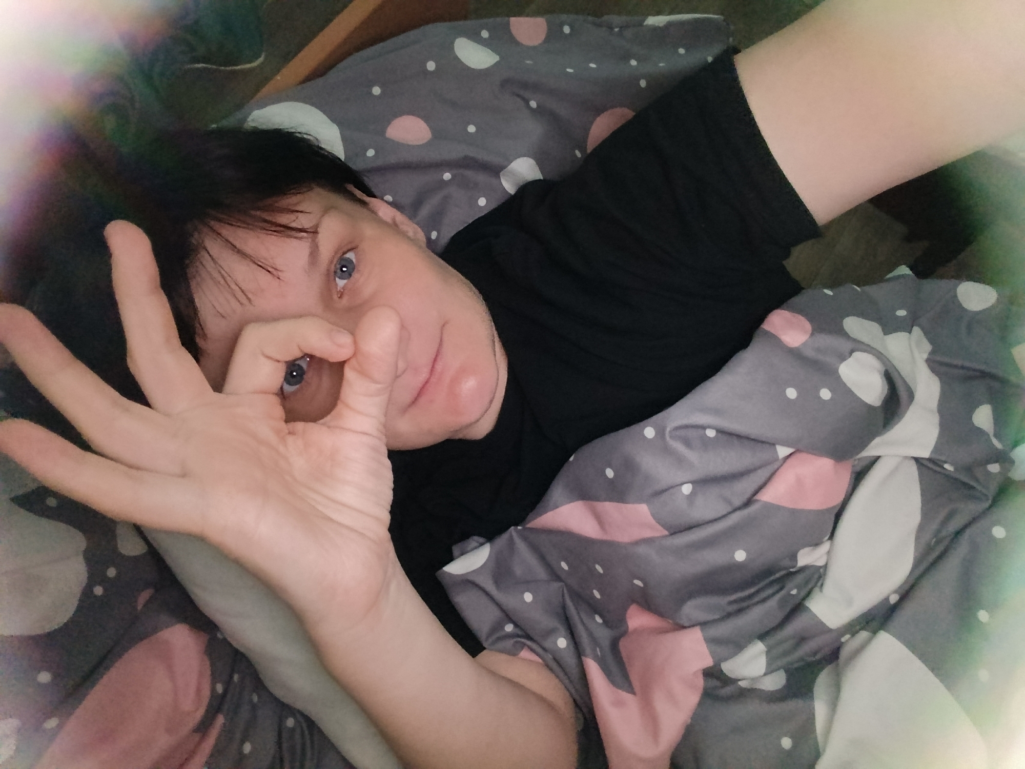 An overhead shot captures a person lying in what appears to be a bed, partially covered by a duvet. The individual, with short dark hair and striking blue eyes, looks directly at the camera. They are wearing a black short-sleeved t-shirt. Their left hand is raised, forming an "OK" gesture with the thumb and index finger, framing their left eye.
The duvet features a modern pattern of abstract grey, pink, and white shapes with small white dots scattered across it. The lighting in the image is soft, with a distinct lens flare on the upper left, suggesting a light source. The person's expression is neutral to slightly soft, contributing to a relaxed mood. The setting appears to be indoors, likely a bedroom, with faint hints of furniture or a wall in the background. No specific location cues indicating "Фоки, Russia" are visible beyond the general domestic environment. There is no visible text in the image.