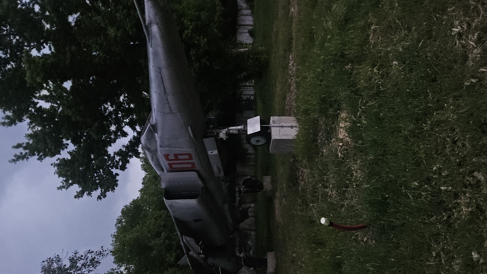 A low-angle outdoor shot shows an aged, gray aircraft on display, possibly a MiG-21, situated on a patch of grass. The aircraft features red numbering "06" near the tail and is supported by concrete blocks. The scene is located within a park or green space, as indicated by the surrounding trees and greenery. A garden hose is visible on the grass. Given the description and the subject matter, it is likely in Plovdiv, Bulgaria, at the Aviation Museum Plovdiv, also known as the Krumovo Air Base.