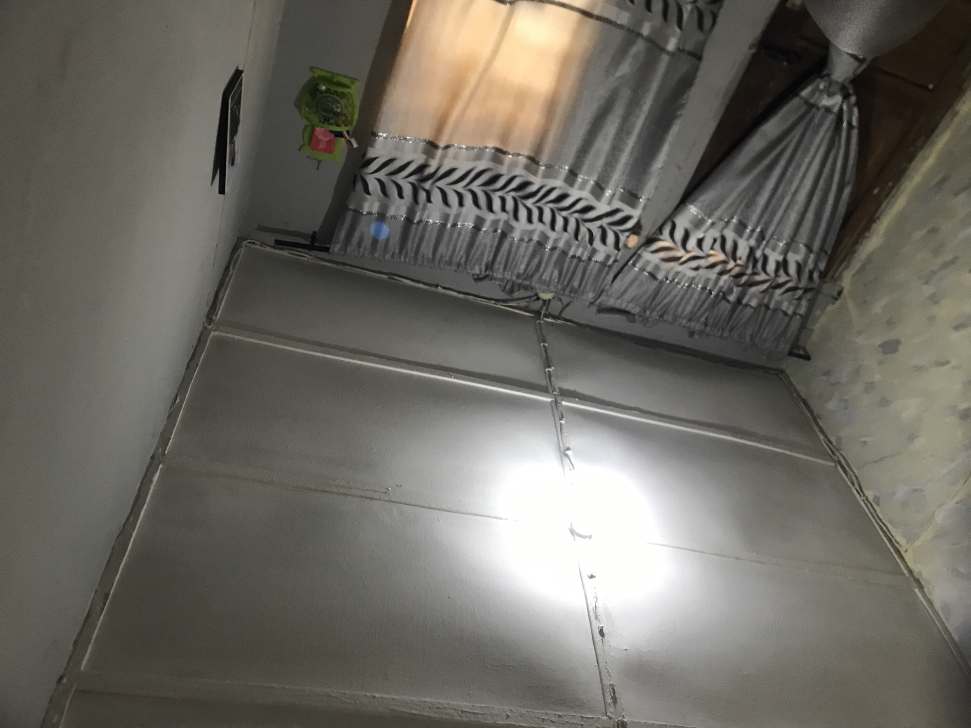This image presents an interior view of a room in Rafin Par, Nigeria. The ceiling is composed of light-colored, possibly plasterboard or sheetrock panels, arranged in a grid pattern with visible seams and surface-mounted wiring. A single, exposed incandescent light bulb is centrally affixed to the ceiling, emitting intense light and creating a significant glare. The wall on the left side of the frame displays a textured, somewhat mottled surface with varying shades, while the right wall is a uniform light gray. Gray curtains with horizontal silver-colored bands and a repeating pattern of black, stylized leaf motifs are drawn across a window or door opening. The bottom portion of the right curtain panel exhibits an orange-yellow illumination, suggesting an external light source. A small, green plastic caddy, possibly an organizer for toiletries, hangs on the right wall, and below it, a dark, rectangular object is mounted vertically. No individuals are depicted in the scene.