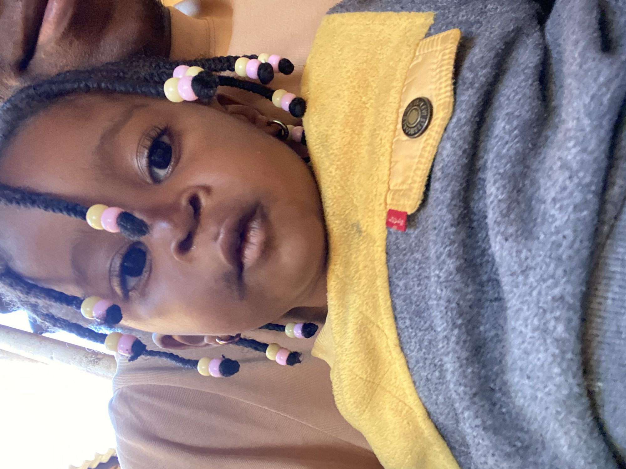 A close-up, eye-level, outdoor portrait shows a young Black child with braided hair adorned with pink and yellow beads. The child is wearing a grey and yellow sweater with a buttoned pocket. The background is blurred, with hints of sunlight and what appears to be a wooden structure. The image is captured in Bukuru, Nigeria.