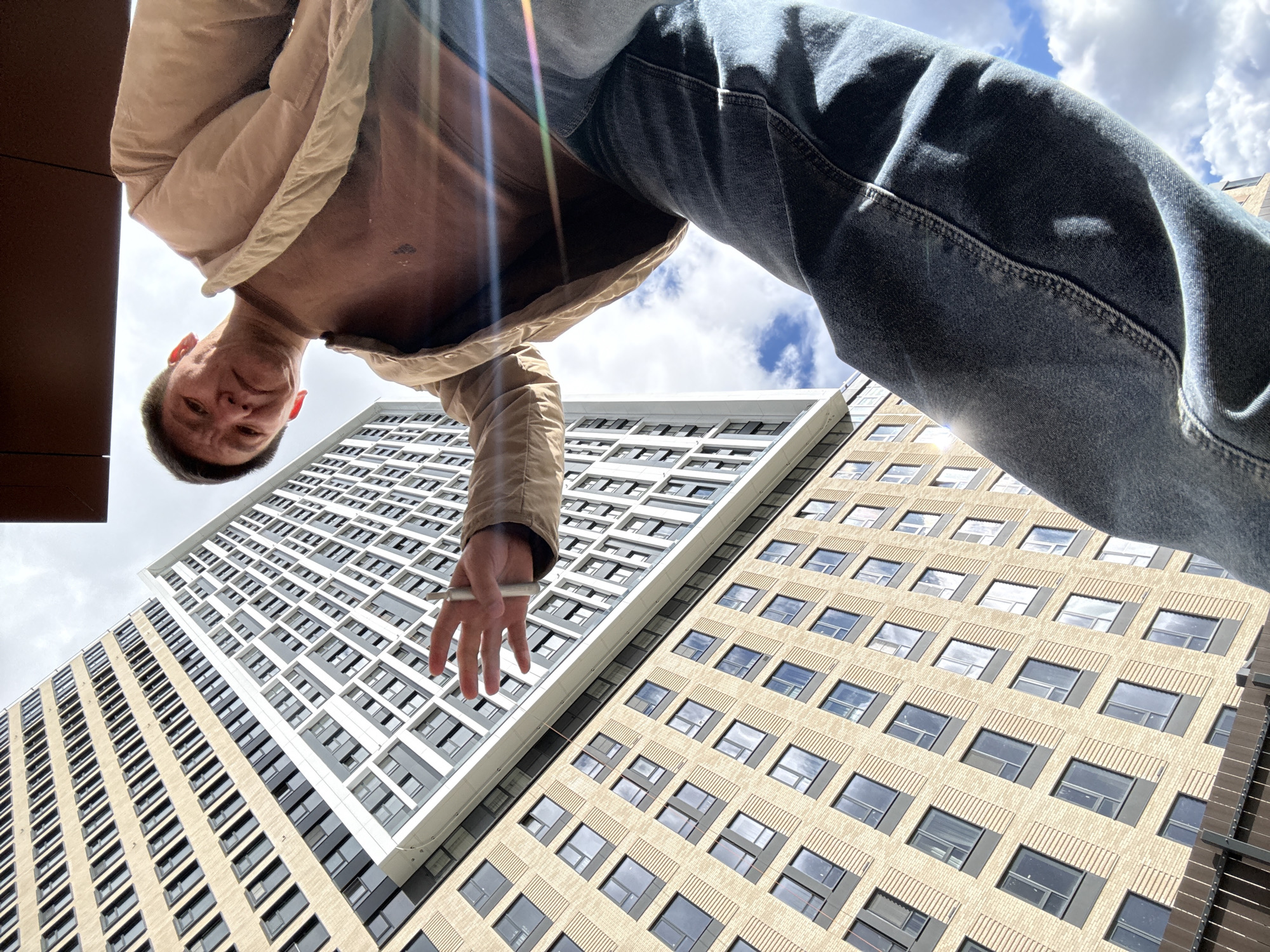 A low-angle perspective photograph shows a man standing above the viewer, with two adjacent buildings in the background. The man is wearing jeans and a light brown jacket open at the chest, and his face is visible. He holds a white object, possibly a cigarette or pen, between his fingers. The buildings are tall, modern structures; one features a white facade with a geometric window design, while the other has a beige brick exterior with repeating window patterns. The sky with some clouds is visible between the buildings and above the man. The photo appears to be taken in the city of Khimki, Russia.