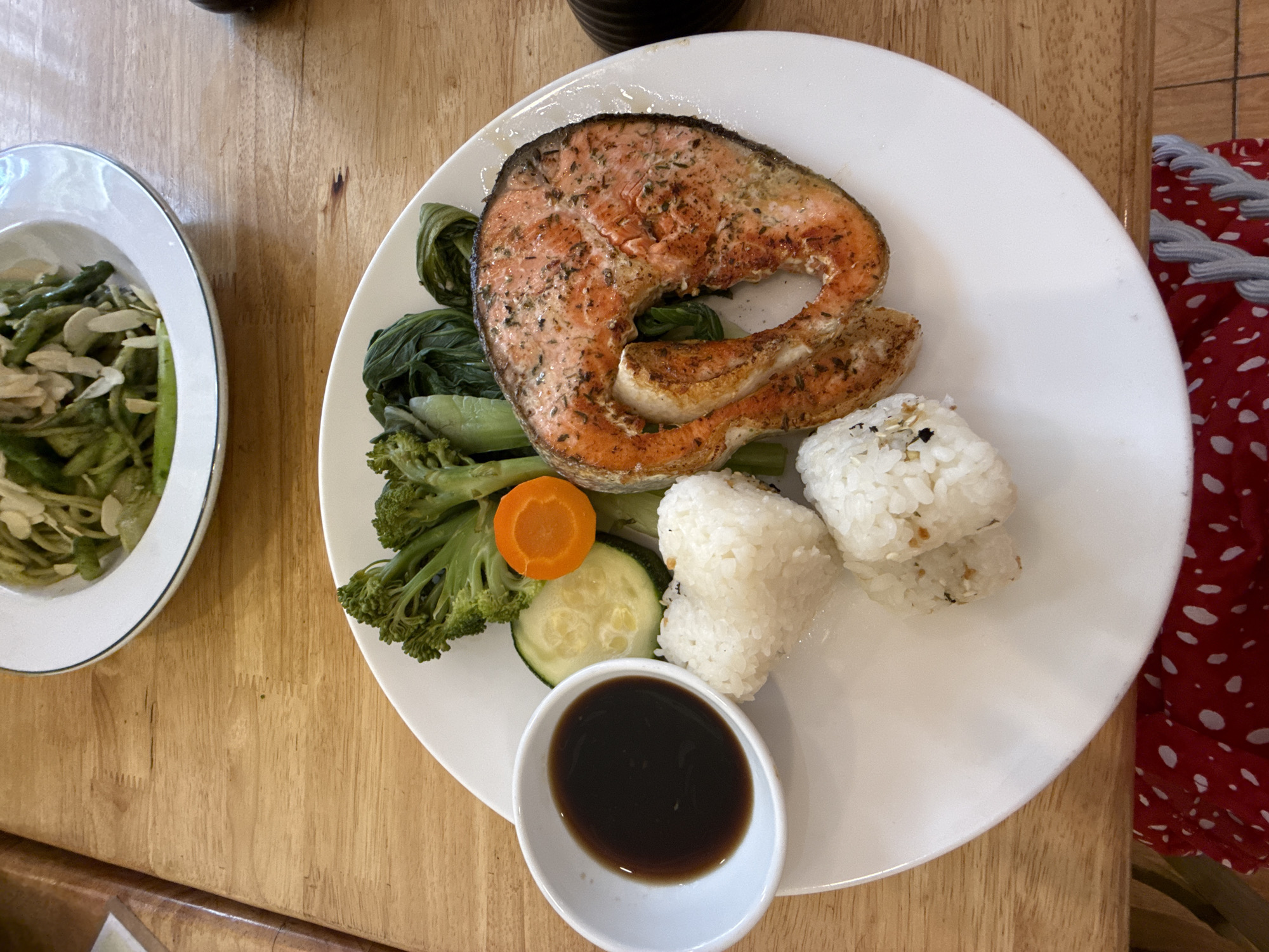 Captured from an overhead perspective, the scene unfolds indoors on a wooden table, displaying a plate of food, another bowl of food, and a glimpse of a garment. The main plate features a grilled salmon steak, accompanied by a mix of vegetables: broccoli florets, a carrot slice, and a zucchini slice. Adjacent to the salmon are two rice balls. A small, oval-shaped bowl contains a dark sauce. To the side, another bowl holds green noodles and slivered almonds. A red and white garment is partially visible at the right edge of the image. The setting likely corresponds to Da Lat, Vietnam, although this could not be forensically determined from image contents.