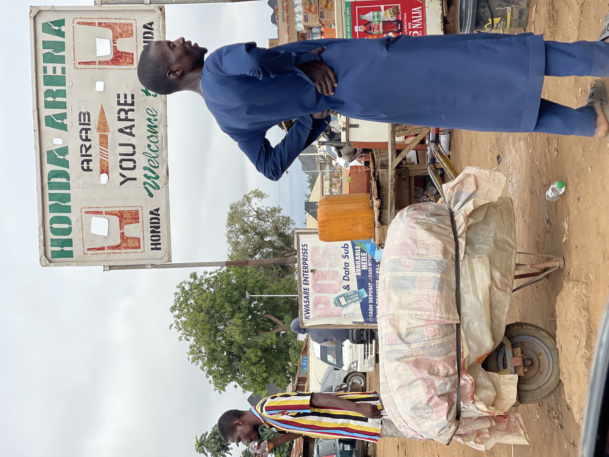 The image depicts an outdoor scene in Abuja, Nigeria, with a prominent sign for "HONDA ARENA" and "ARAB YOU ARE Welcome to NIDA". In the foreground, a man wearing a blue traditional outfit stands with his hands on his hips, facing to the right. To his left, another man in a colorful striped shirt is drinking from a plastic cup. A wheelbarrow loaded with a large, white, tarp-covered bundle is positioned in the center. Behind the wheelbarrow, various market stalls and vehicles are visible, including a white van. The ground appears to be dirt and gravel. The sky is overcast.