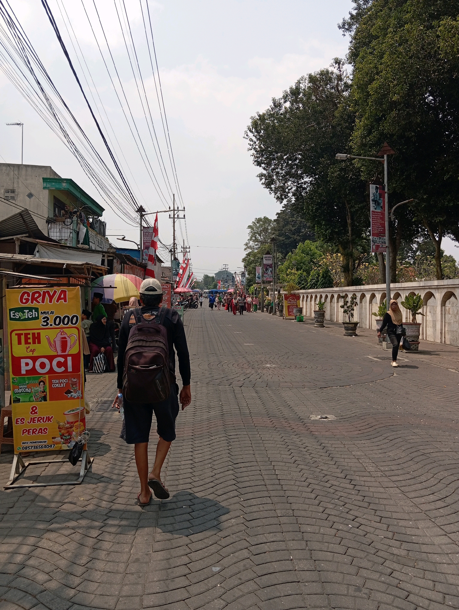In Blitar, Indonesia, a street scene unfolds with a man walking away from the camera, sporting a backpack. To his left, a yellow food stall prominently displays a sign for "Griya Es Teh," advertising various tea drinks. The stall features images of tea cups and other beverages, along with prices and contact information. The street itself is paved with interlocking bricks, some of which appear to be unevenly laid, creating a wavy pattern. On the right side of the street, a woman in a hijab walks past a row of arched structures and potted plants. Flags and banners are visible along the street, suggesting a festive or commemorative occasion. Buildings and trees line the background, and power lines crisscross the sky. The overall atmosphere suggests a bustling urban environment.
