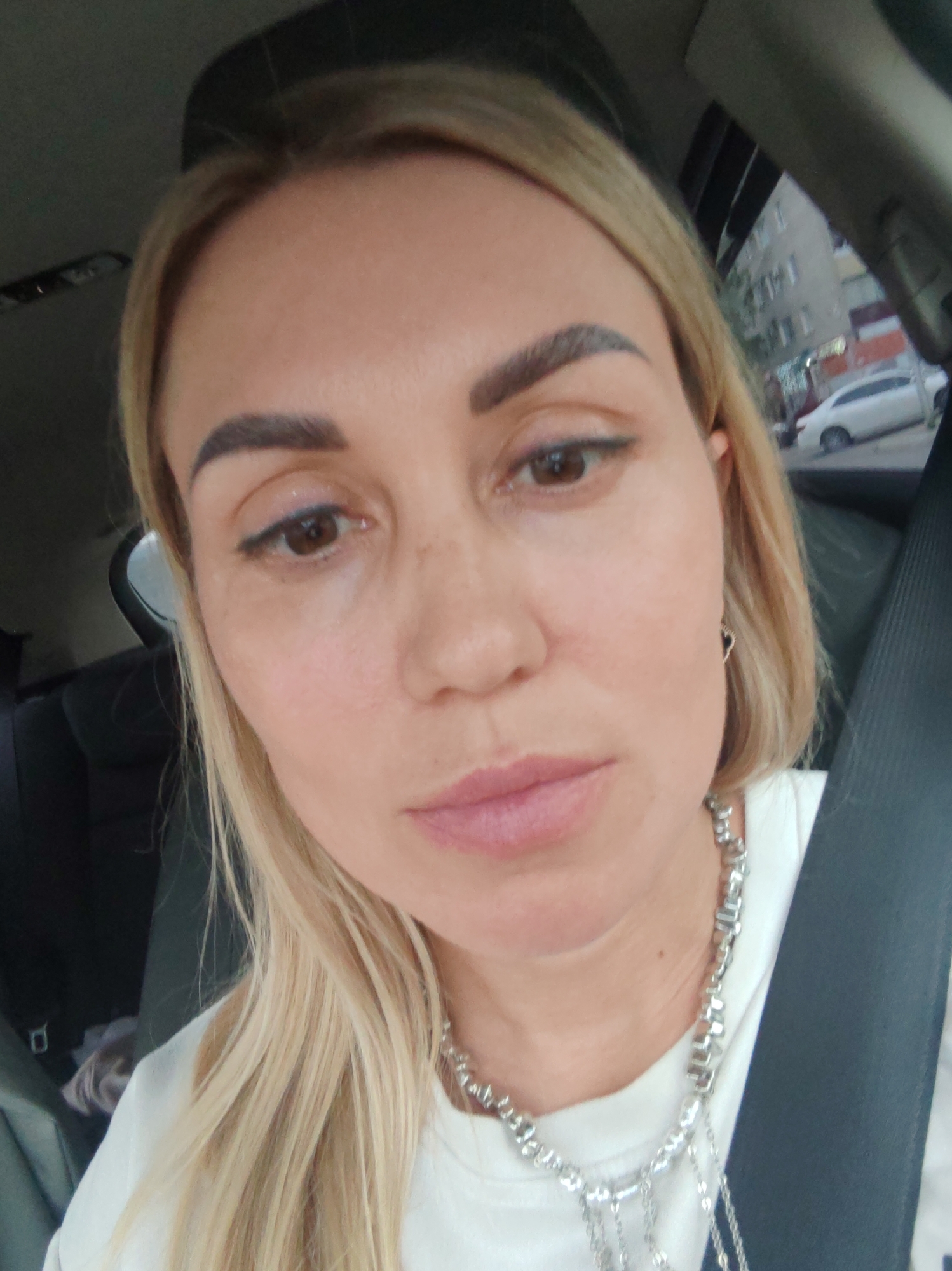 A woman with blonde hair and fair skin, wearing a white shirt and a silver necklace, is sitting in the passenger seat of a car. Her gaze is directed towards the camera, and her expression is neutral. The car interior is visible, including a black seatbelt and the headrest of the driver's seat. Outside the car window, a street scene in Komsomolsk-on-Amur, Russia, can be observed, featuring buildings, a white car, and parked vehicles. The lighting suggests it is daytime, with overcast skies contributing to a diffused natural light.
