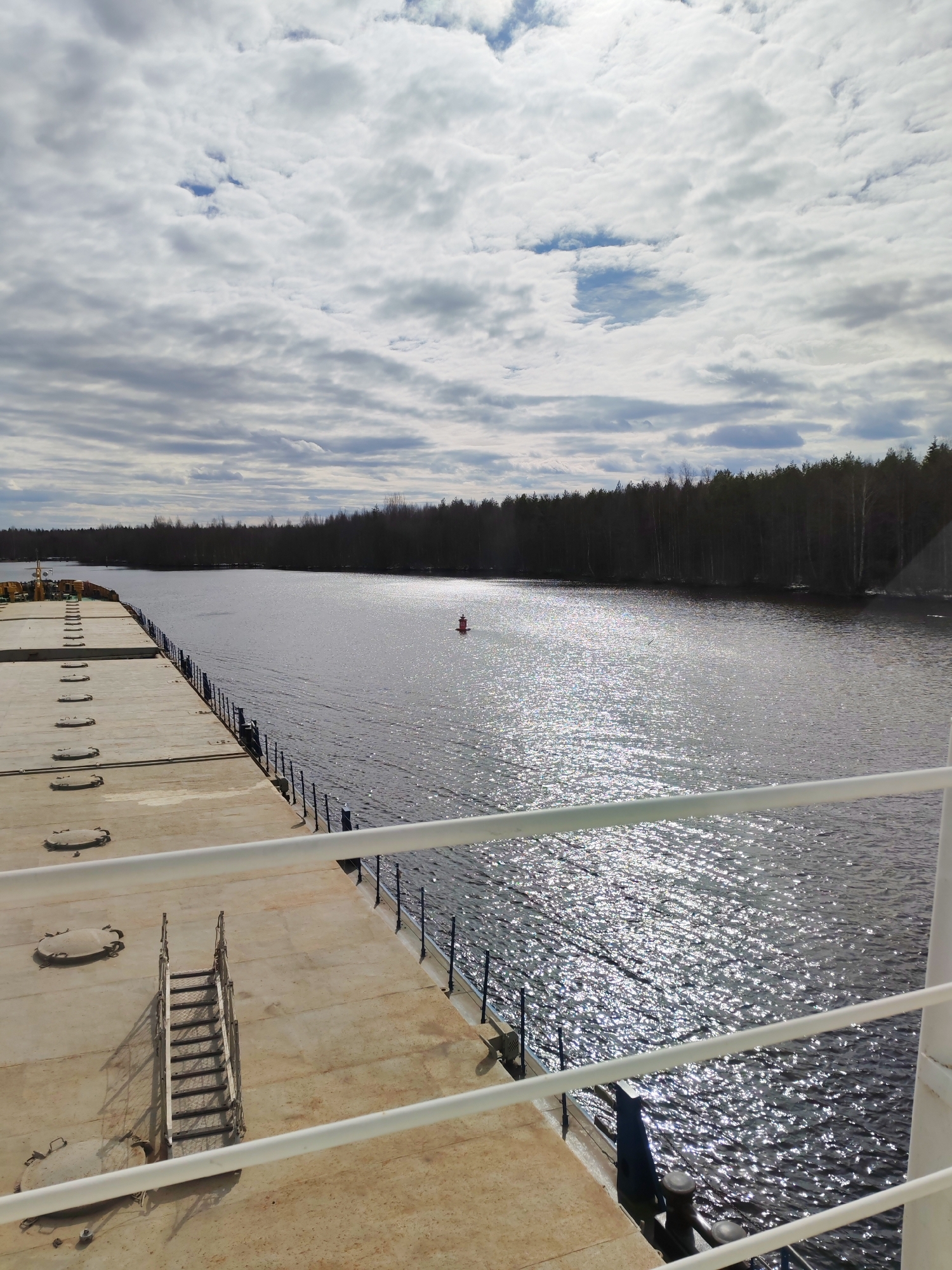 The image captures a wide inland waterway, likely a river or canal, viewed from the deck of a large vessel, possibly a cargo ship or barge. In the foreground, the ship's expansive, flat deck is visible, featuring several oval-shaped hatch covers and a portable metal staircase. White railings run along the side of the deck, partially framing the scene.
Beyond the ship, the dark water shimmers intensely with sunlight reflections, suggesting a bright day despite the cloudy sky. A red, conical navigational buoy floats in the mid-distance on the water, indicating a marker in the channel. The far bank of the waterway is lined with a dense, dark forest of deciduous trees, mostly bare, implying either early spring or late autumn. The tree line extends far into the background, following the course of the water.
The sky above is predominantly covered with light grey and white stratocumulus clouds, with occasional patches of blue sky visible through breaks in the cloud cover. The overall atmosphere is calm and expansive. No people or visible text are present in the image. The scene suggests a vessel navigating or moored in a natural or semi-natural environment, characteristic of an inland waterway in Kurdiug, Russia.