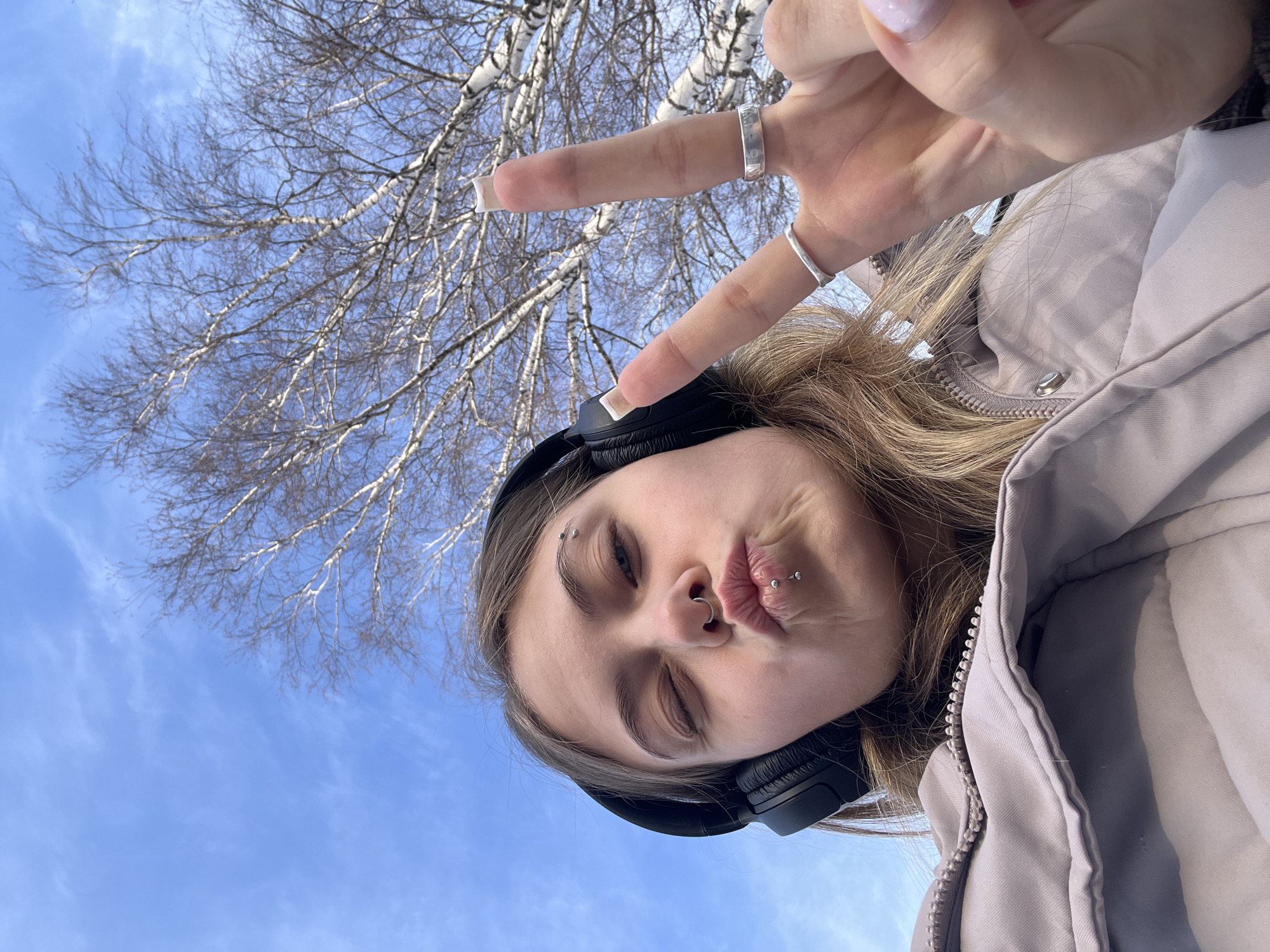 An individual is depicted in a low-angle selfie, positioned against a clear blue sky with sparse, wispy white clouds and bare deciduous trees, indicative of late winter or early spring in Фоки, Russia. The subject, a young female with blonde hair, wears black over-ear headphones and a light-colored, possibly lavender or grey, padded zippered jacket. Her right eye is closed in a wink, and her lips are pursed in a pouting expression. Visible facial piercings include a curved barbell on her left eyebrow, a hoop in her septum, and two studs on her lower lip. Her left hand is raised, forming a peace sign with the index and middle fingers. This hand displays two silver-toned rings on the ring finger and middle finger, and her fingernails have light-colored polish.
