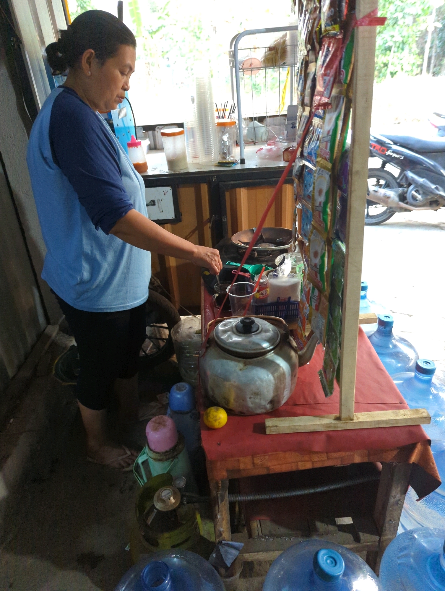 A medium-shot, eye-level, outdoor, daylight image shows a woman in Kedungdowo, Indonesia, standing behind a small makeshift stall. She is wearing a blue t-shirt with navy blue sleeves and black leggings. Her hair is pulled back in a bun. The woman is in the process of preparing something, holding a small glass in her right hand, and a spoon in her left hand, with a kettle and other items on the counter in front of her. The stall is set up on a dirt ground with some debris.
Behind the woman, the stall features various items for sale, including bags of snacks and drinks. A motorcycle is parked to the right of the stall. Large water jugs are visible in the foreground and background, suggesting the stall may also sell water. A propane tank and other containers are also present. The scene appears to be in a casual, possibly residential, setting.