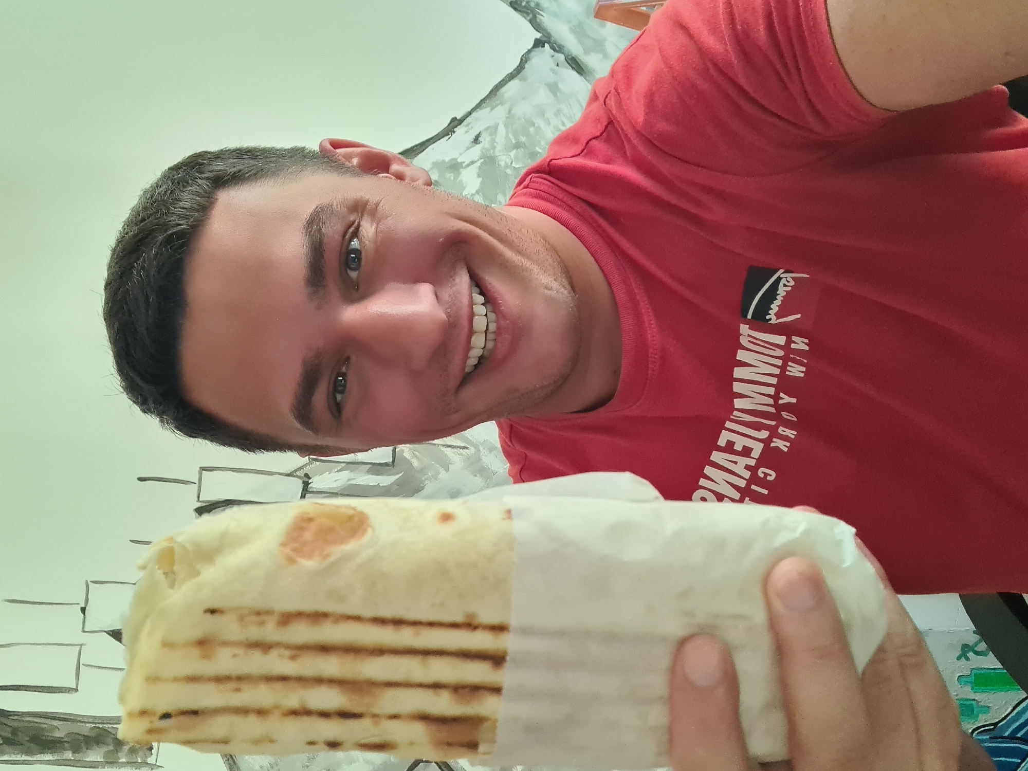 A man with short dark hair and blue eyes is smiling and holding a wrap in his right hand. He is wearing a red T-shirt with white lettering. The wrap is toasted and partially covered in white paper. The background consists of a wall with a drawn design resembling a brick wall and mountain scene. The location in the city of Budva, Montenegro is not discernible from the image.