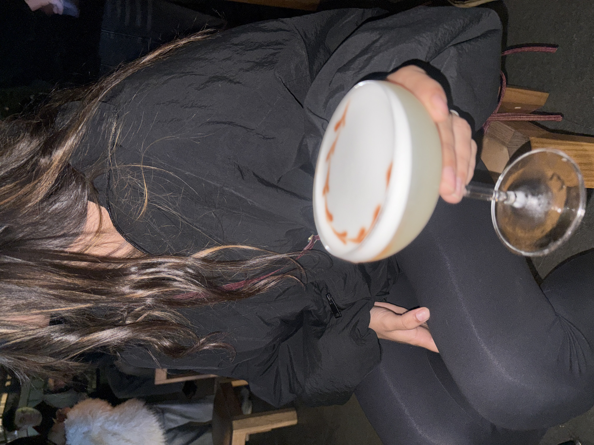 A person with long, brown hair sits wearing black leggings and a black puffy jacket. She is holding a cocktail in a stemmed glass. The cocktail has a white foam top with a decorative design of brown liquid on top. A partial view of a wooden bench is visible. There is no clear indication of the location being in Buenos Aires, Argentina.