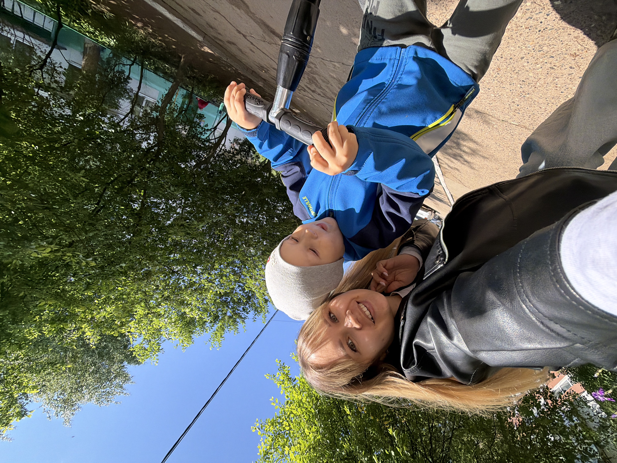 This is a brightly lit outdoor selfie taken on a sunny day, featuring a woman and a young child in Chaikovsky, Russia. The image is taken from a low angle, looking upwards towards the subjects and the clear blue sky.
The woman, likely taking the photo, is positioned in the foreground on the right. She has long blonde hair, fair skin, and is smiling directly at the camera. She is wearing a black leather-like jacket over a white top.
To her left and slightly behind her is a young boy, who appears to be a child. He wears a light grey beanie, a blue and dark blue jacket with yellow accents, and dark grey pants. He is holding the handlebars of a scooter, which has black grips and a silver stem, and is looking upwards past the camera.
The background features lush green trees with abundant foliage, suggesting a season with full leaves. Patches of bright blue sky are visible between the tree canopy. A thin power line stretches across the sky. The ground below appears to be a paved surface, likely concrete or asphalt. A distant building is partially visible through the trees in the lower left portion of the image.
Visible text includes "Scooter" on the scooter's black handlebar grip, and "ON" and "OFF" indicators on the scooter's stem mechanism. The child's jacket also has partially visible, reversed text that reads "NORTHVILLE" near the zipper. The overall mood is cheerful and bright, indicated by the woman's smile and the clear, sunny weather.