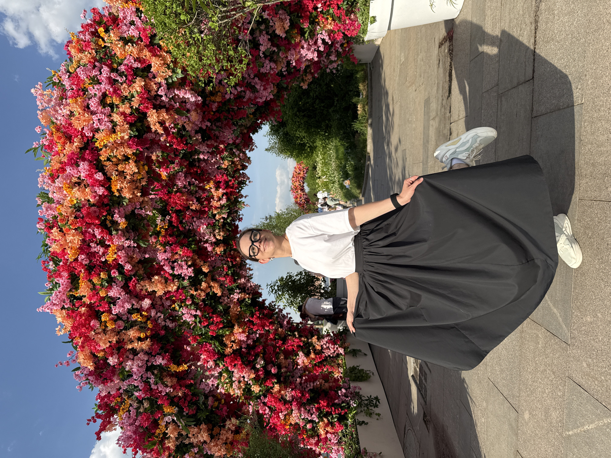 A woman is standing under a floral arch in what appears to be a public park in Moscow, Russia. The floral arch is constructed of many red, orange, and pink flowers. The woman is wearing a white t-shirt, a black skirt, and glasses. She is smiling and looking at the camera. The background includes trees, other park visitors, and pavement. The weather appears sunny.