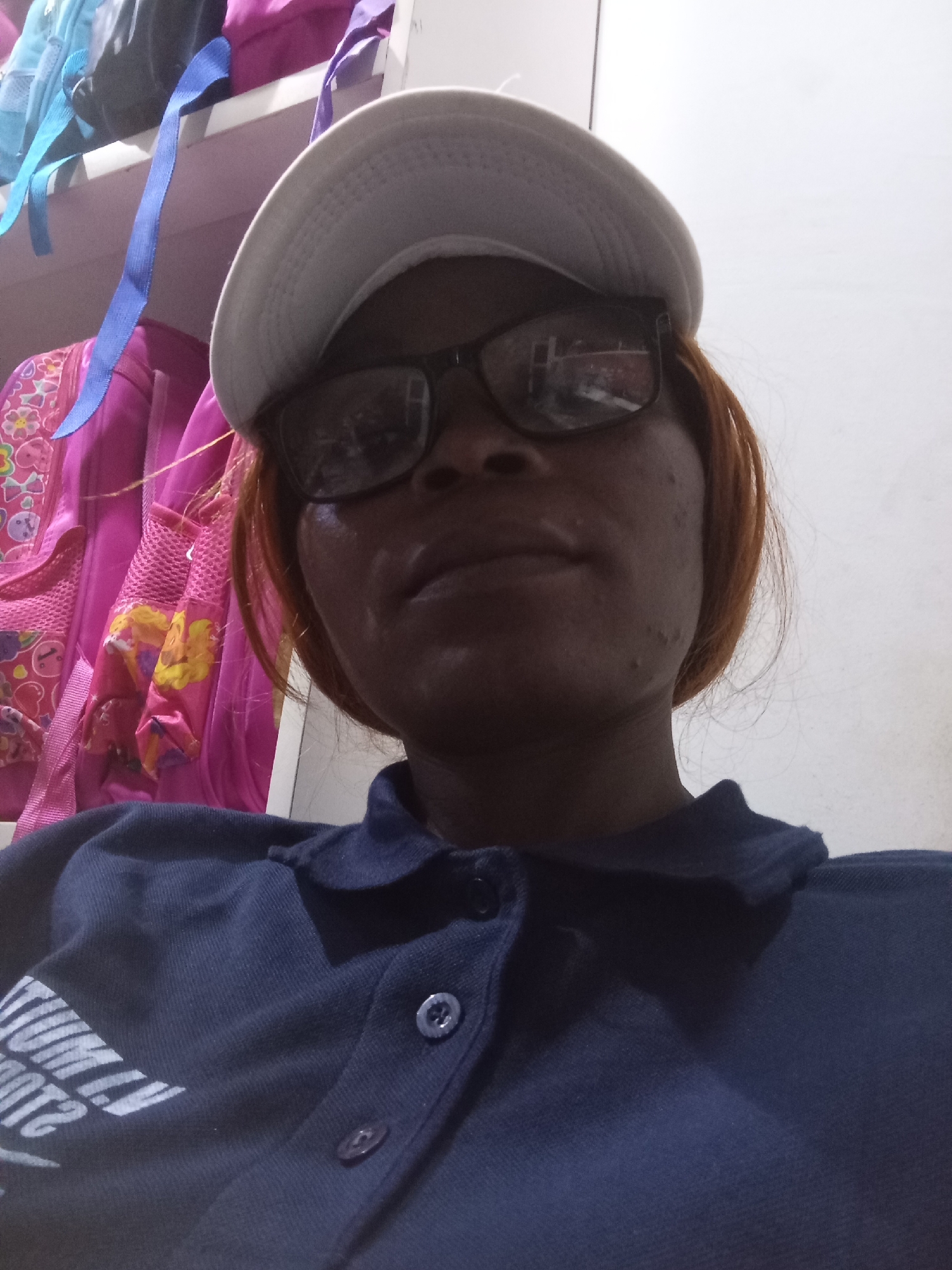 A woman wearing a white baseball cap and dark-rimmed glasses is captured in a close-up selfie. She has reddish-brown hair and is wearing a navy blue polo shirt with white text on the left side of the chest. The background is cluttered with several backpacks, mostly in shades of pink and blue, suggesting a storage area or a room with multiple bags. The lighting is somewhat dim, casting shadows on the woman's face and the surrounding objects. The scene appears to be indoors, possibly in a residential setting. The specific location in Howayi, Nigeria, is not discernible from the visual information provided in the image.