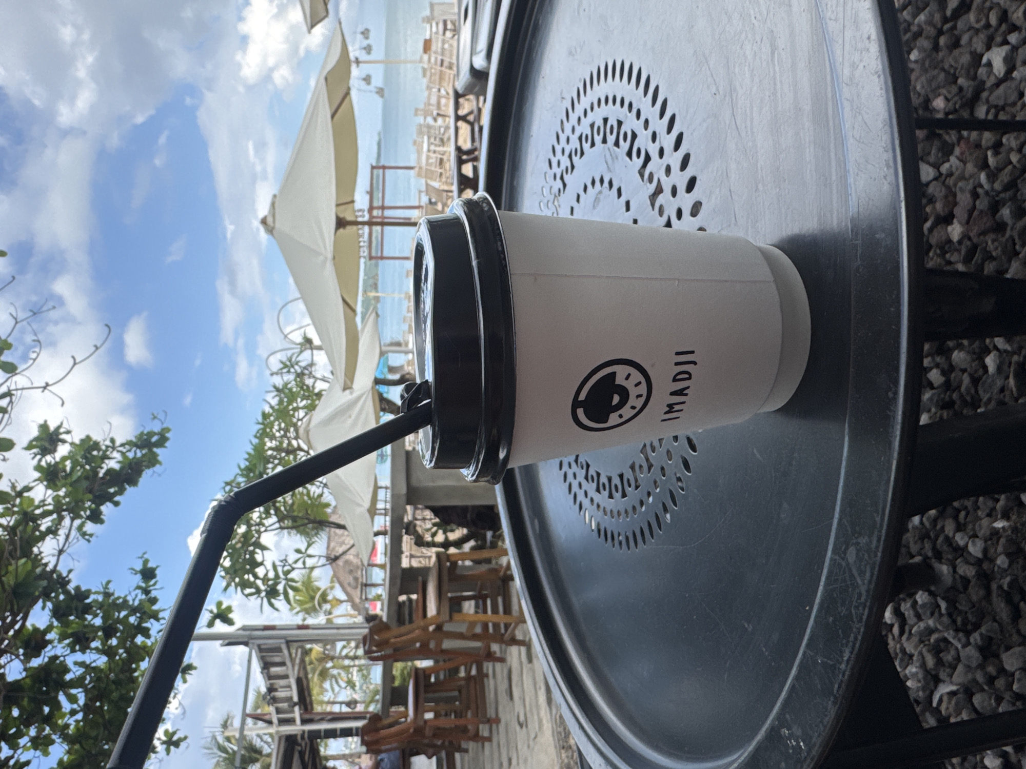 A close-up, medium-shot image captures a disposable white coffee cup with a black lid and straw, resting on a small, round, perforated black table outdoors. The cup features the logo "IMADJI" on its side.
In the background, out-of-focus elements reveal an outdoor dining area with wooden chairs and tables under large white parasols. Beyond this area, the ocean and a partly cloudy sky are visible. Green trees and vegetation are seen on the left side of the image. The location is likely Kuta, Indonesia, considering the beachside setting and the branding on the cup, but this is not confirmed with certainty in the image.