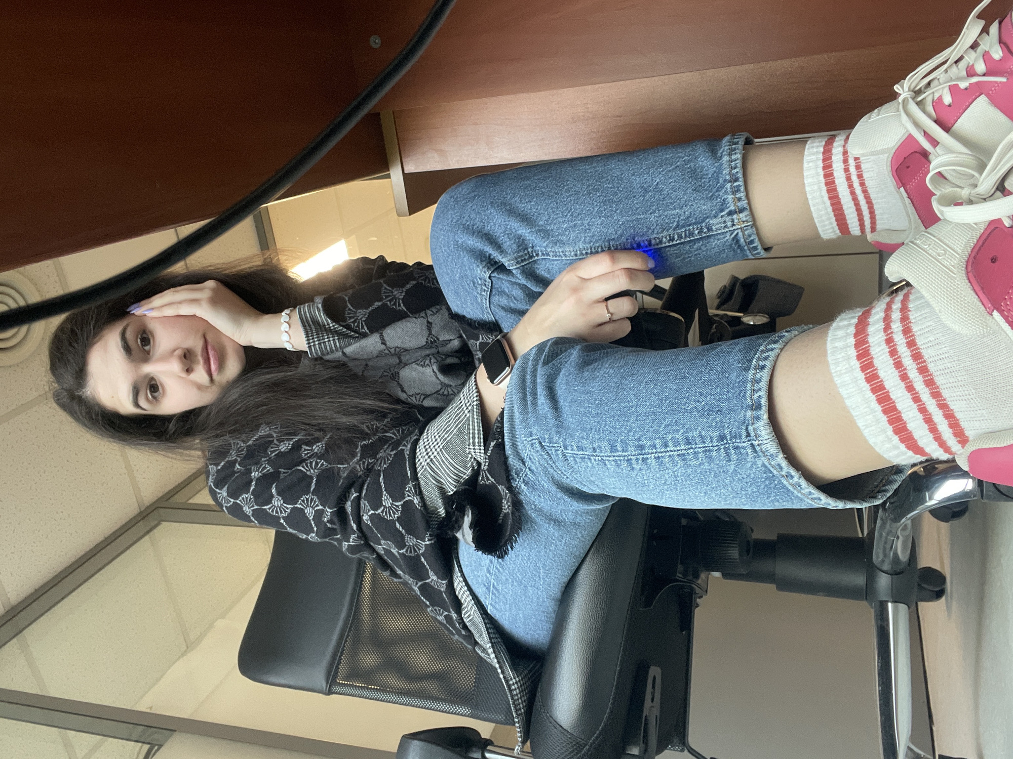 A young woman is sitting in a black office chair beneath a desk. She is wearing blue jeans, white socks with red stripes, and pink and white sneakers. She is also wearing a patterned black and white jacket. She is holding a black object with a blue light in one hand. She is wearing a watch on her wrist. The location appears to be an office setting. The city of Sofia, Bulgaria, cannot be determined from the image.