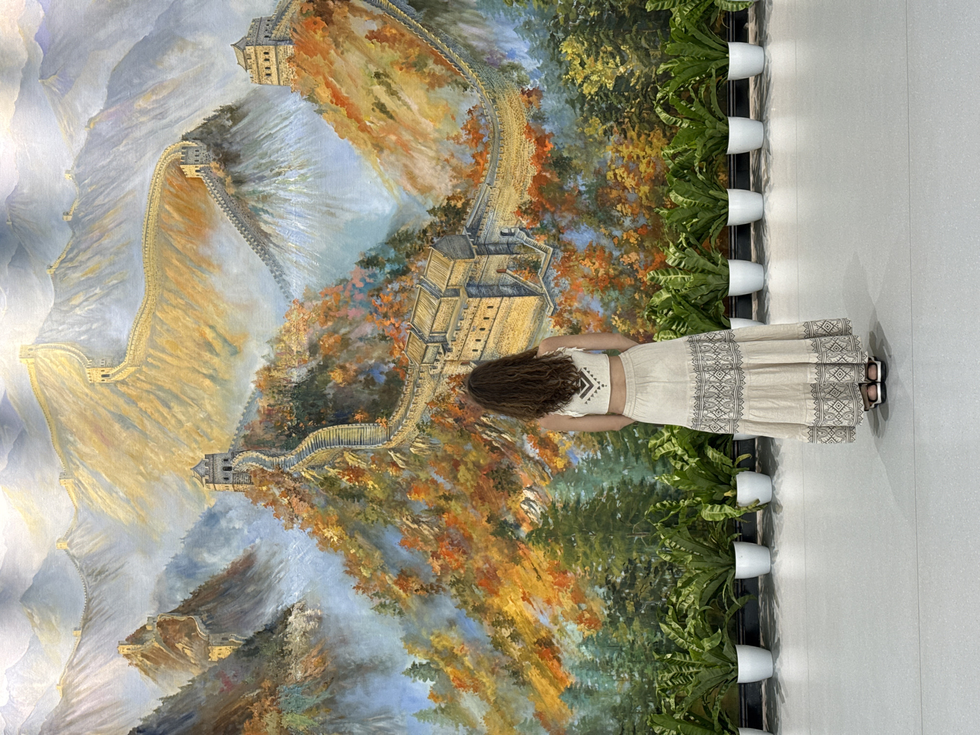 A woman with curly brown hair stands facing an oil painting of the Great Wall of China. She is wearing a white crop top and a long patterned beige skirt with sandals. Her back is visible. The painting is detailed, showing the wall snaking through mountains and foliage in autumnal colors. To the right of the painting, a row of ferns is planted in white pots along a black ledge. The floor is a light-colored, polished tile. The setting is indoors and the location is Huairou District, China.