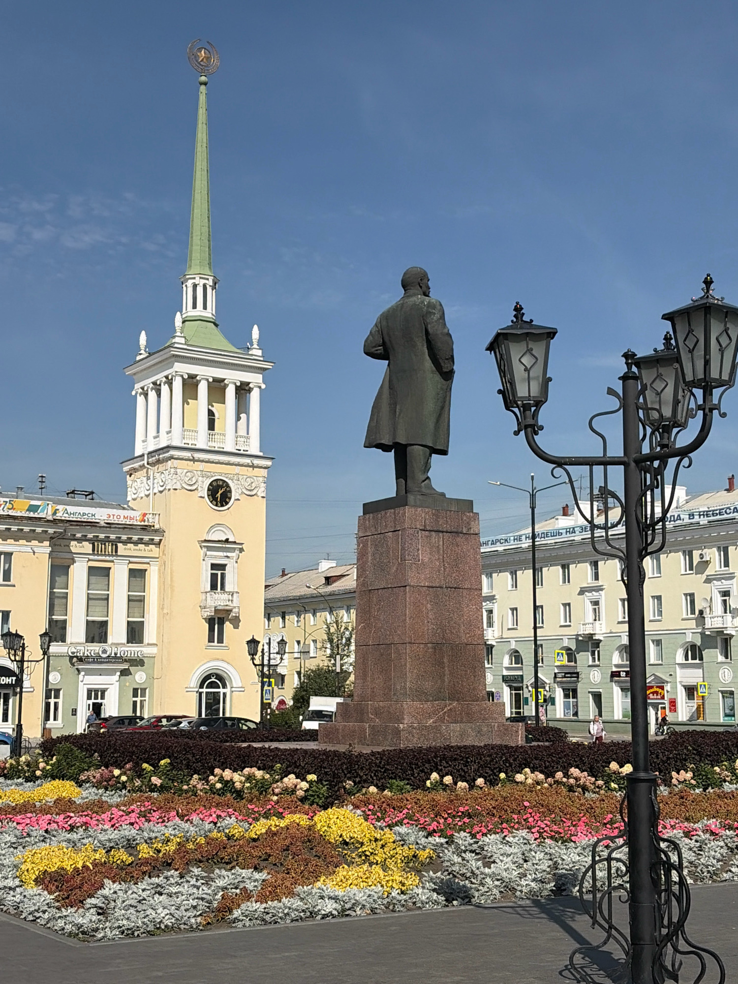 The image captures a public square in Angarsk, Russia, on a clear day. Dominating the foreground is a statue of Vladimir Lenin, facing away from the viewer, positioned atop a granite pedestal. To the left of the statue is a tall, yellow building with a green spire topped by a Soviet-era star and hammer and sickle emblem. This building features a large clock face. In front of the buildings and the statue, a vibrant flower bed showcases a colorful arrangement of plants, including yellow, pink, and white varieties, bordered by dark foliage. Several ornate street lamps with multiple lights stand in the foreground and mid-ground, casting shadows on the paved square. In the background, multi-story buildings with light green and yellow facades line the square. A few people are visible in the mid-ground, walking or standing, and several cars are parked near the buildings. The overall scene depicts a typical urban environment with historical monuments and civic landscaping.