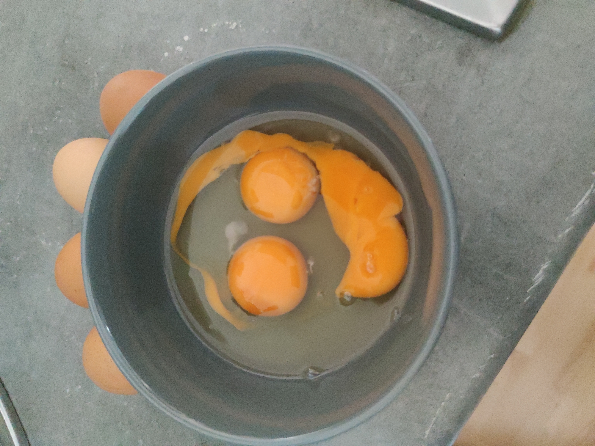 A top-down shot shows a gray bowl containing three cracked eggs with bright orange yolks and transparent egg whites. Several uncracked brown eggs sit beside the bowl. The bowl is on a gray countertop next to a stainless steel object and a light-colored wooden surface. There are no people visible in the scene. There is no indication that this image was taken in Barcelona, Spain.