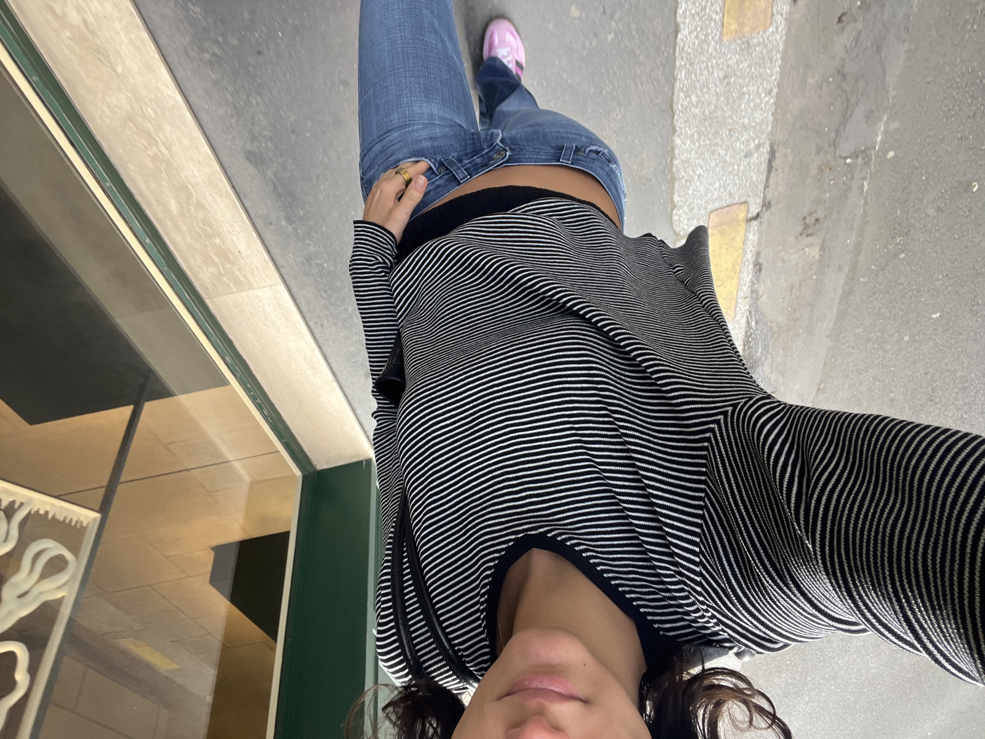 A close-up, downward-facing shot captures a person from the chest to the feet, walking or standing on an urban sidewalk. The person wears light blue jeans, a black and white horizontal striped long-sleeve top, and bright pink sneakers. A black bralette or undershirt is visible beneath the striped top, peeking above the waistline of the jeans. A prominent gold-colored ring adorns the left hand.
The ground consists of varied textures, including light gray concrete, darker asphalt, and sections of lighter-toned, possibly tiled, pavement. To the left, a building facade is visible, featuring a dark green frame around a large reflective glass panel. Reflections in the glass show an interior ceiling with square tiles and a white, possibly decorative, object. To the right, the pavement includes some light yellow or tan rectangular markings. The lighting suggests it is daytime. There is no visible text in the image.